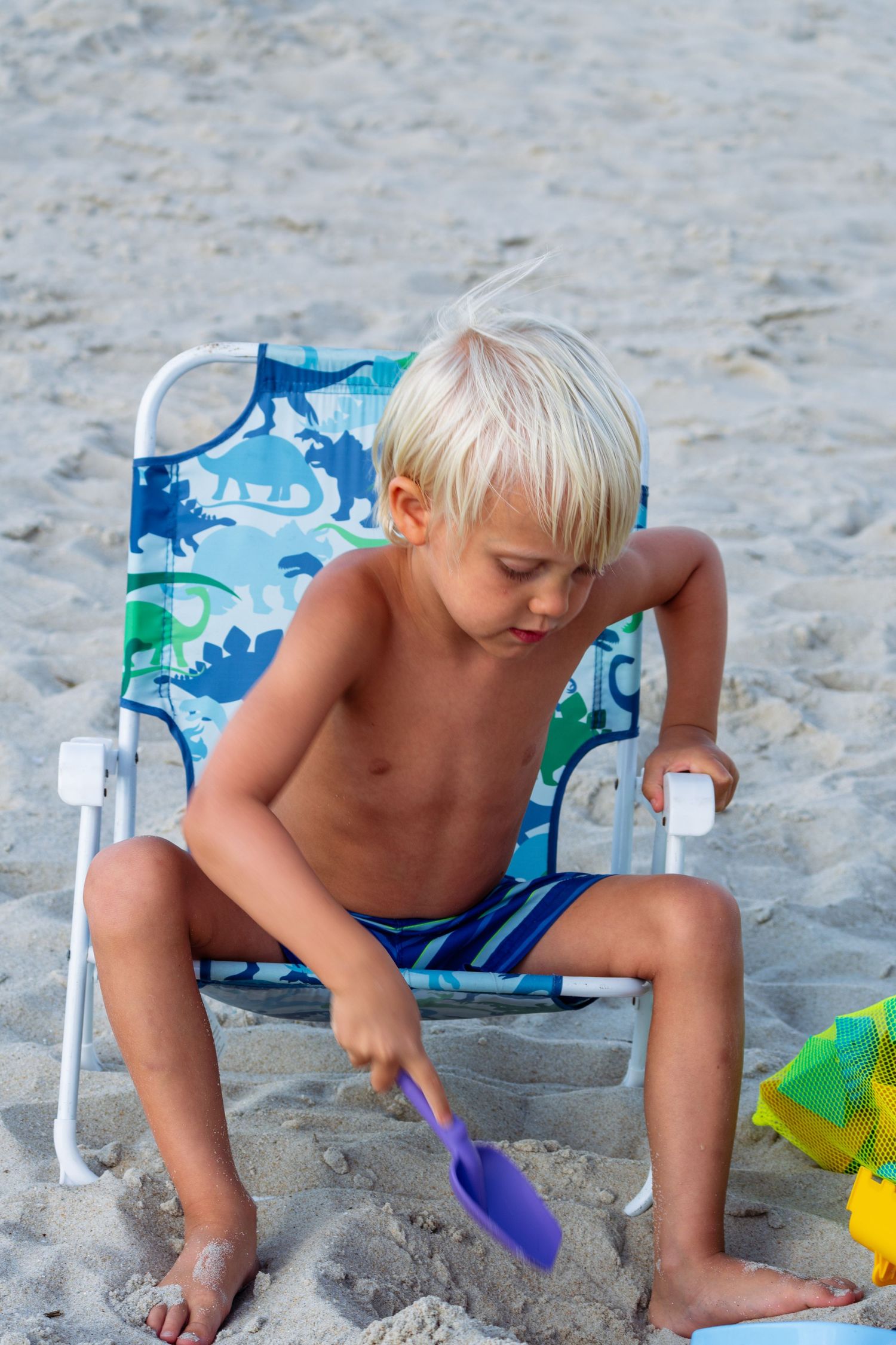 A blonde toddler boy sitting in a small beach chair playing with a purple shovel in the sand.