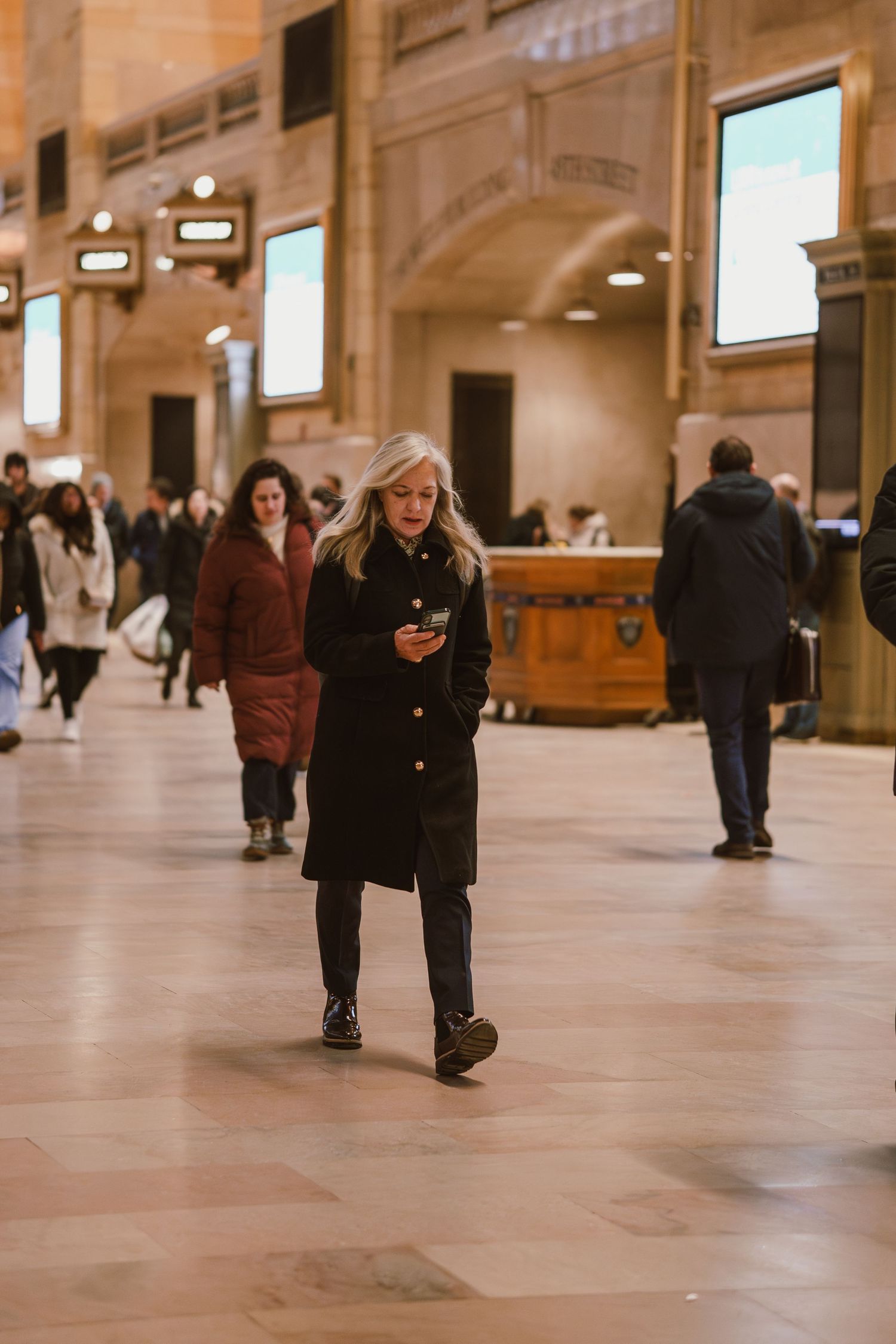 A blonde woman in a black coat checks her phone while walking through Grand Central Terminal.