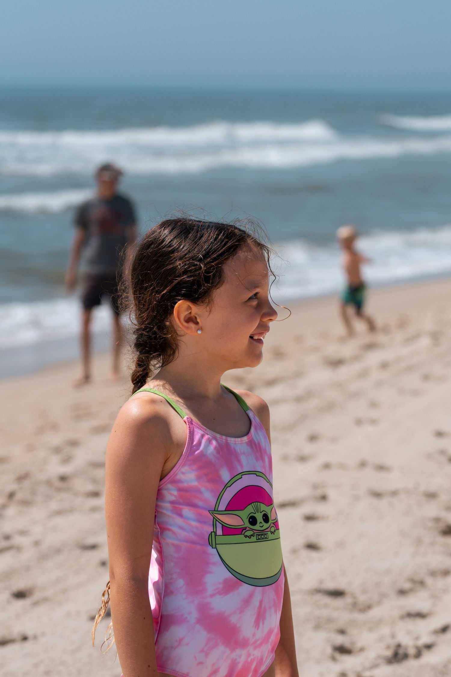 A young girl in a tie-dye Baby Yoda swimsuit smiles on a sunny beach with waves and people in the background.