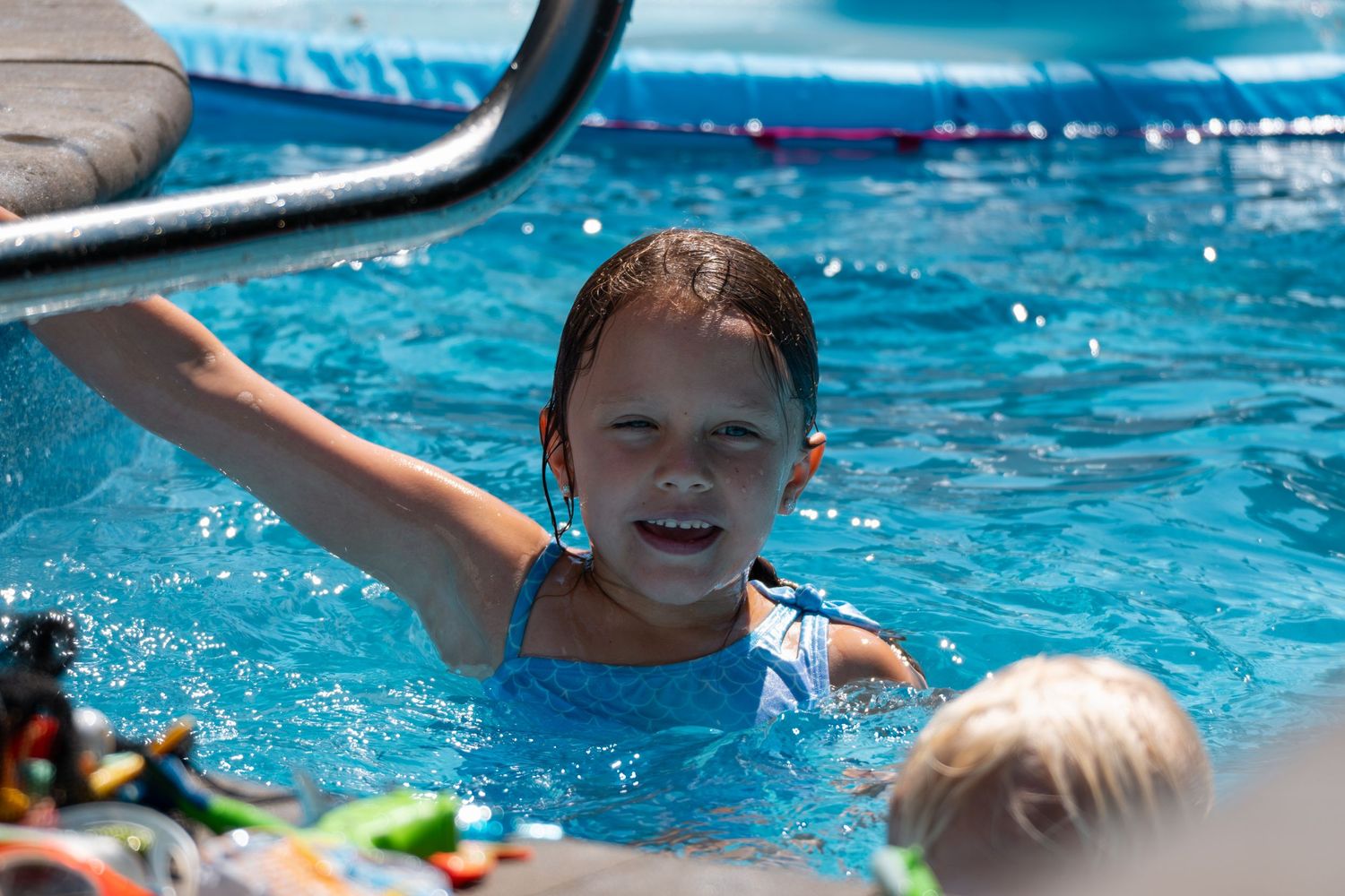 Cheerful girl in blue swimsuit waving arm while swimming in a bright outdoor pool.