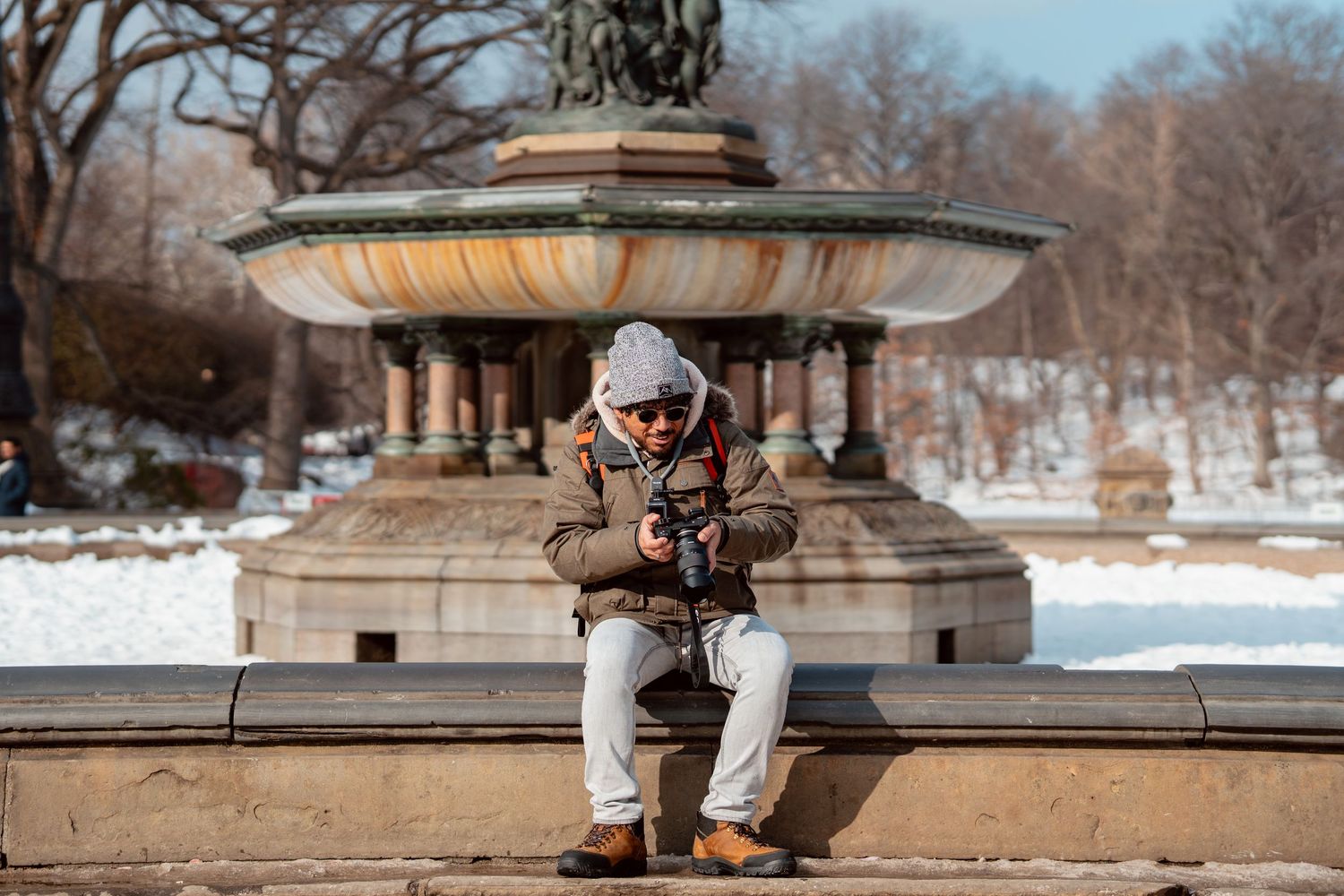 Male photographer reviewing camera in front of Bethesda Fountain in snowy Central Park winter scene.