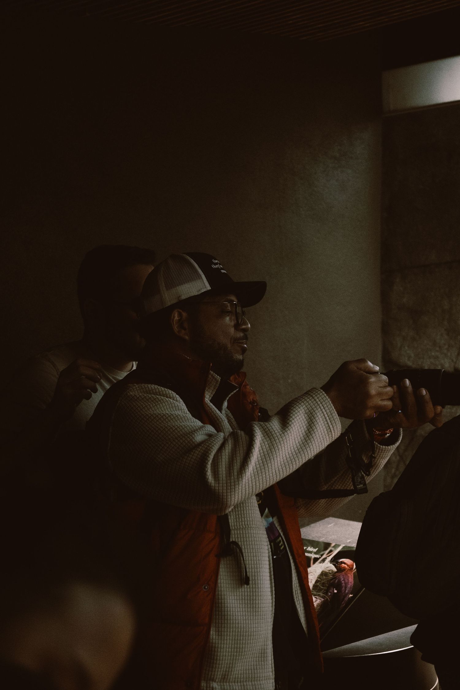 A man wearing a trucker cap holding a camera in a dimly lit indoor setting.
