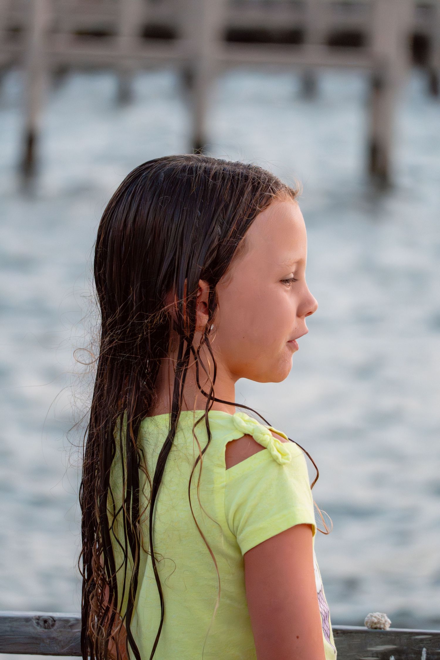 Profile portrait of a young girl with wet dark hair wearing a yellow shirt near the water.