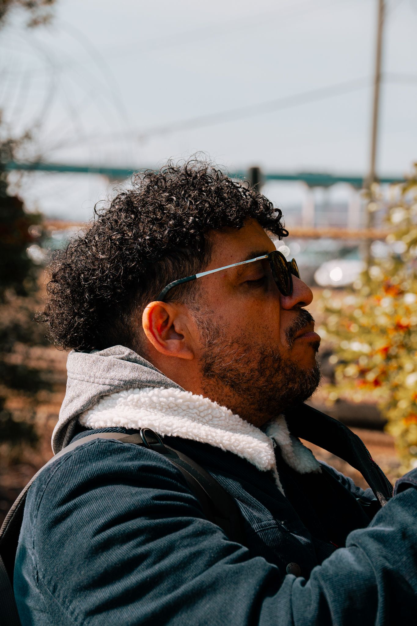Man with curly hair and sunglasses gazing thoughtfully outdoors with a bridge in background.