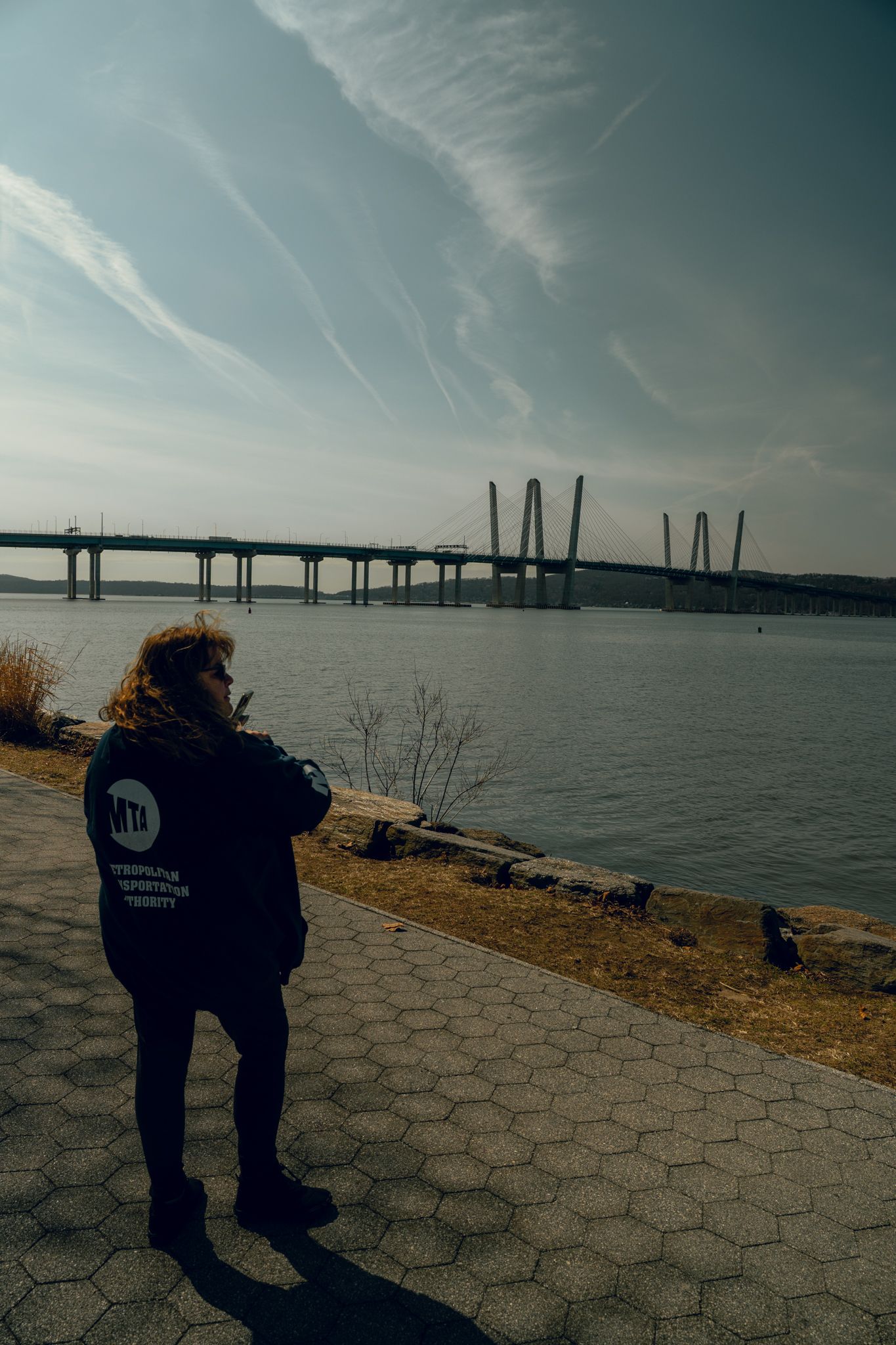 Person in MTA jacket stands on a paved riverside path viewing a large cable-stayed bridge under a streaked sky.