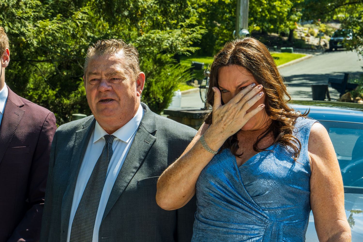 A man in a suit and a woman in blue dress appear emotional outdoors on a sunny day.