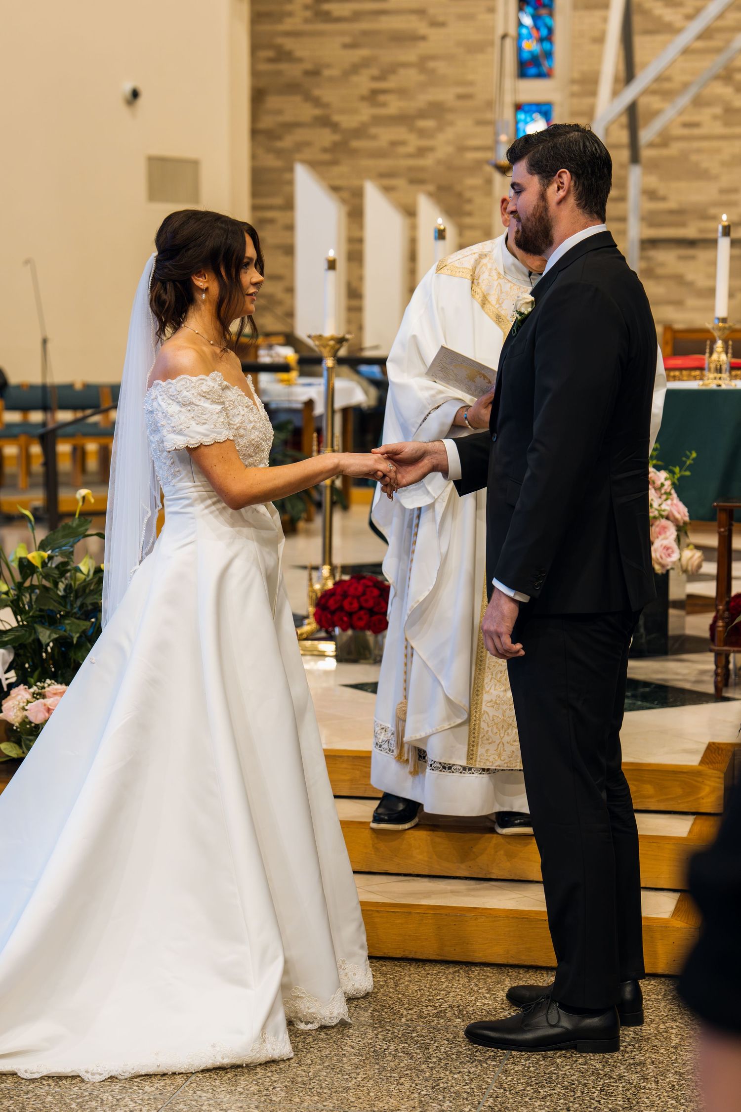 Bride and groom exchange vows at church altar with officiant, bride in white off-shoulder gown with veil.