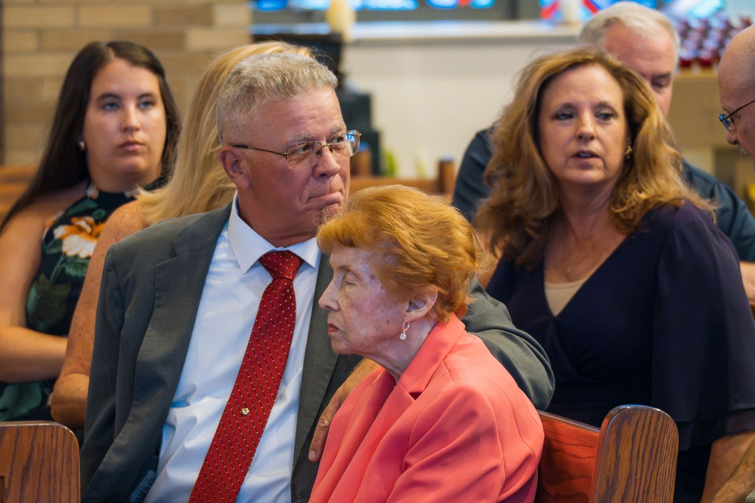 Three adults seated in church pews during a formal gathering, including an elderly woman in pink and a man in a red tie.
