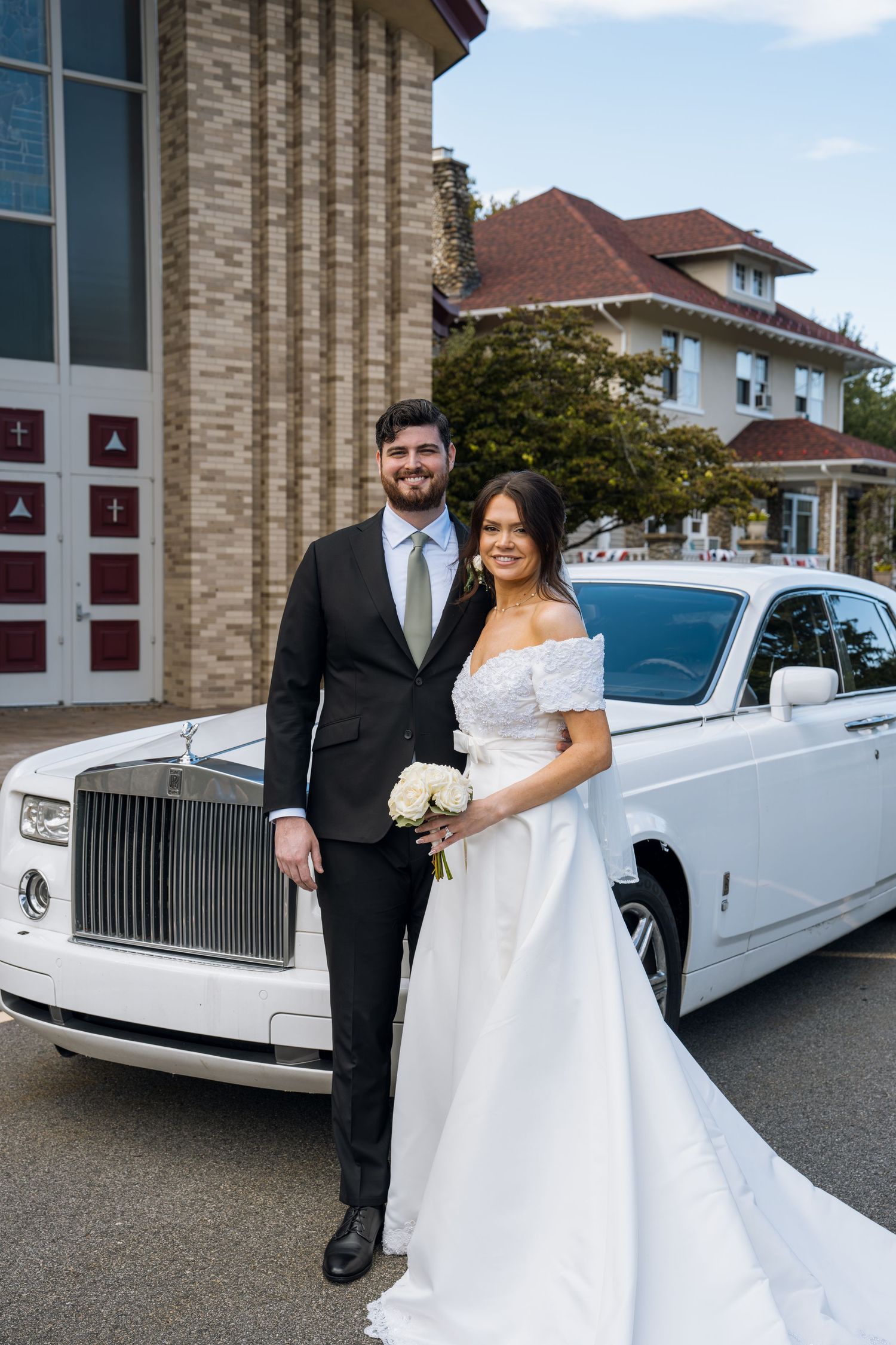 Bride and groom pose in front of a white Rolls-Royce outside a church on their wedding day.