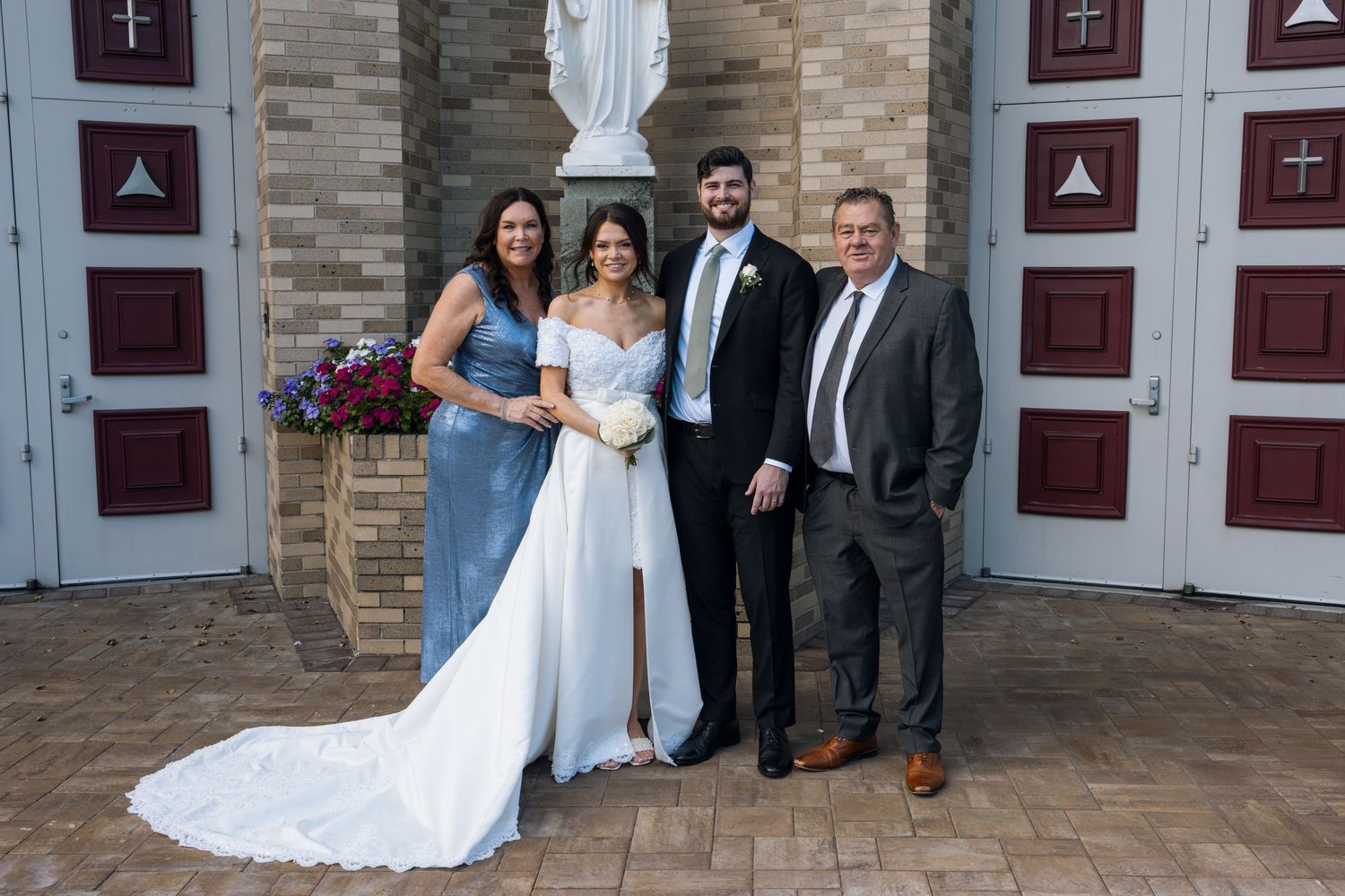 Wedding party poses outside church, bride in white gown with train, groom in suit, flanked by family members.