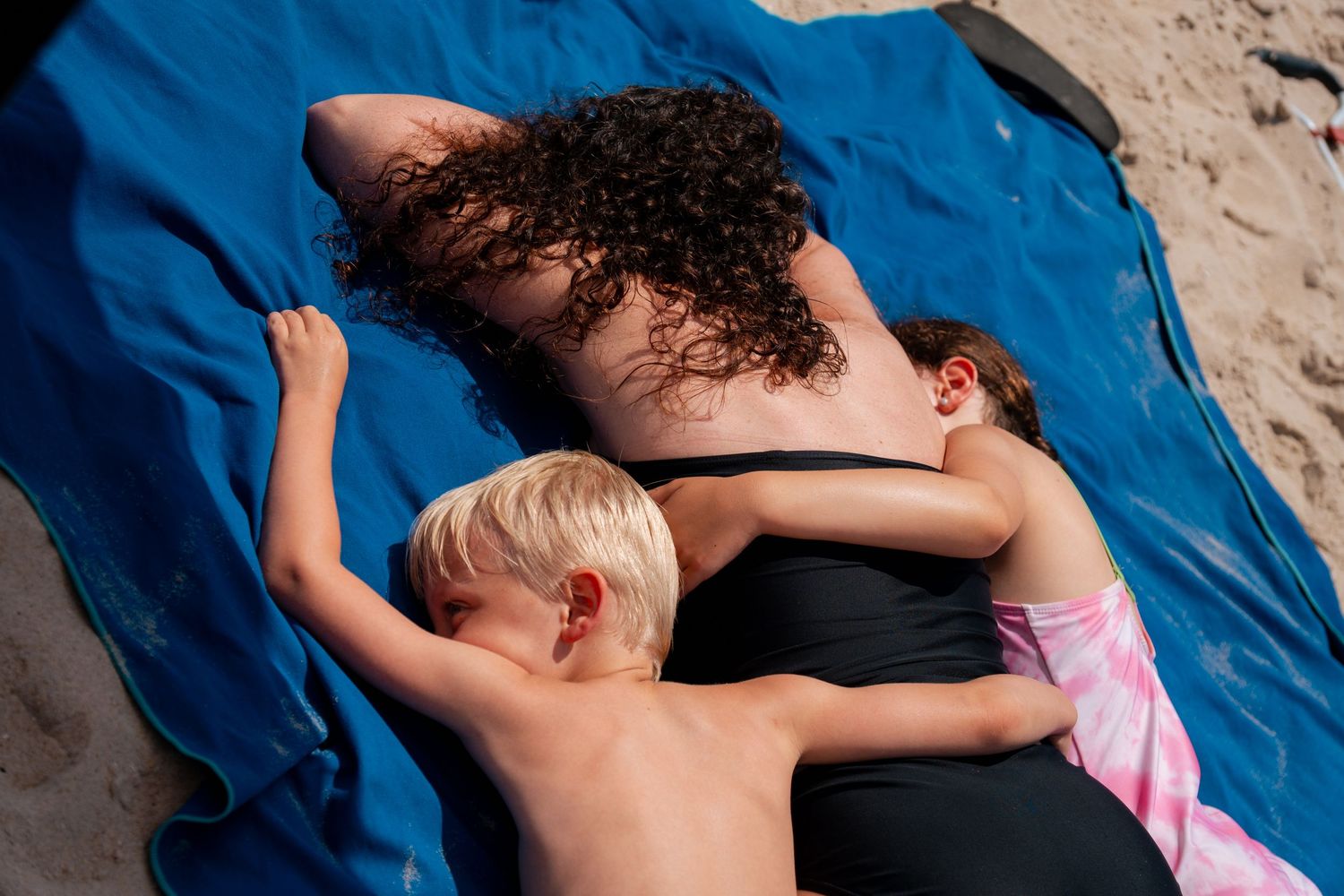 Woman in black swimsuit lying on blue towel at beach with two young children cuddling beside her.