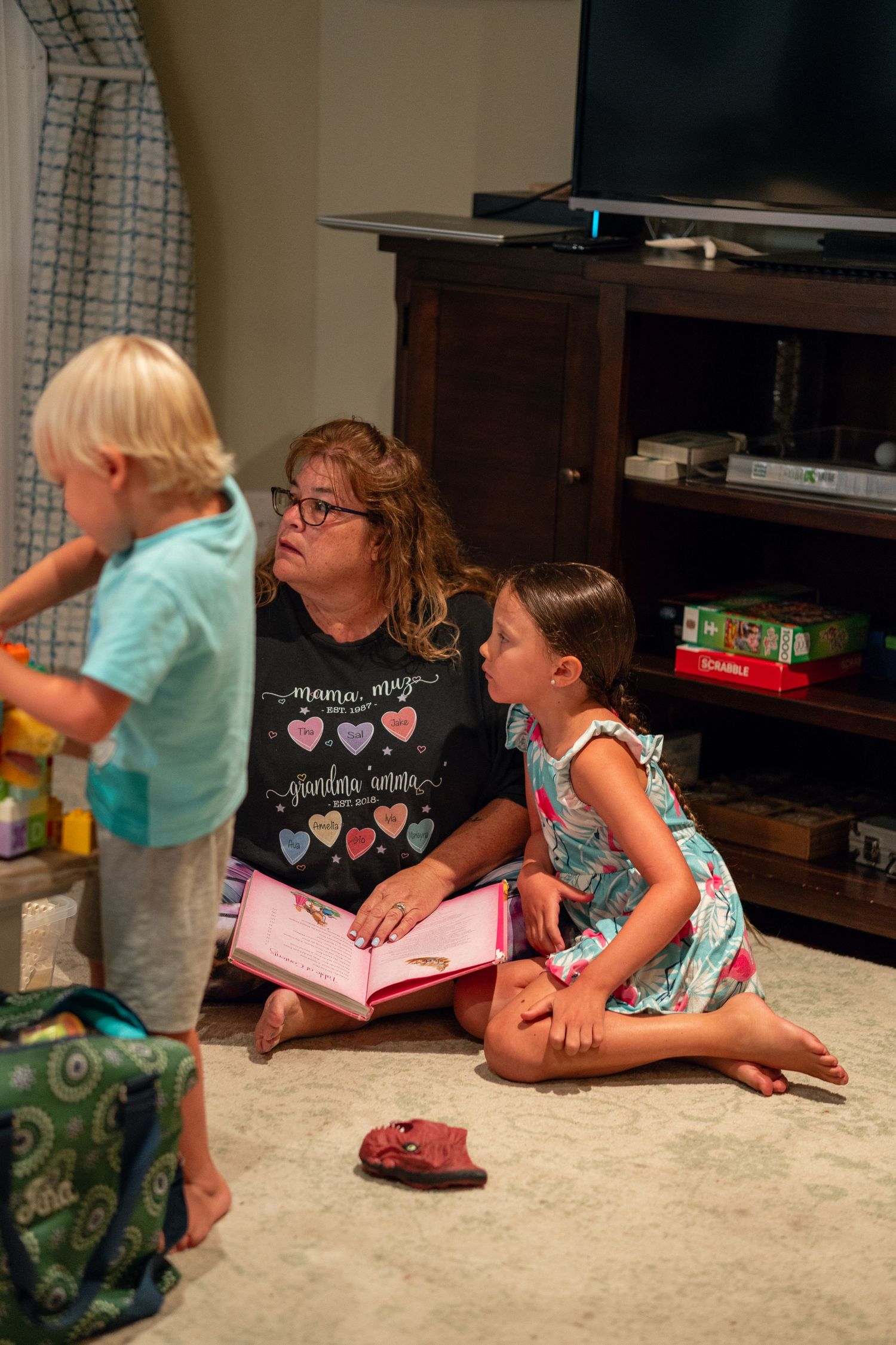 Woman sitting on floor reading a book with a young girl near a TV stand indoors.