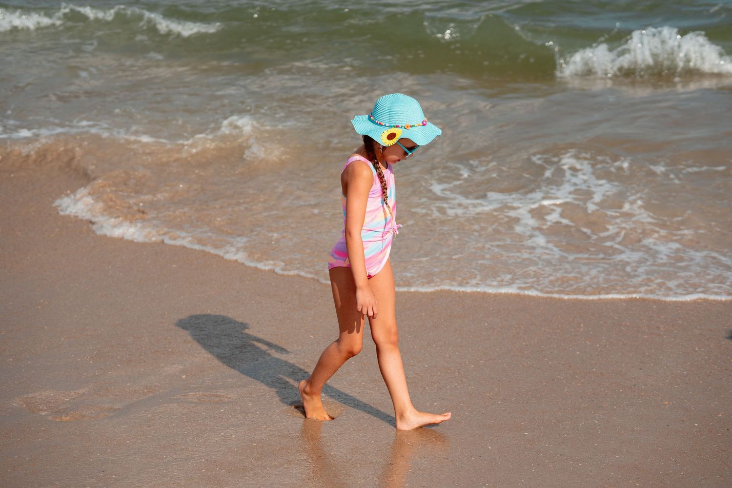Young girl in blue sun hat and pink swimsuit walking along a sunny ocean shoreline.