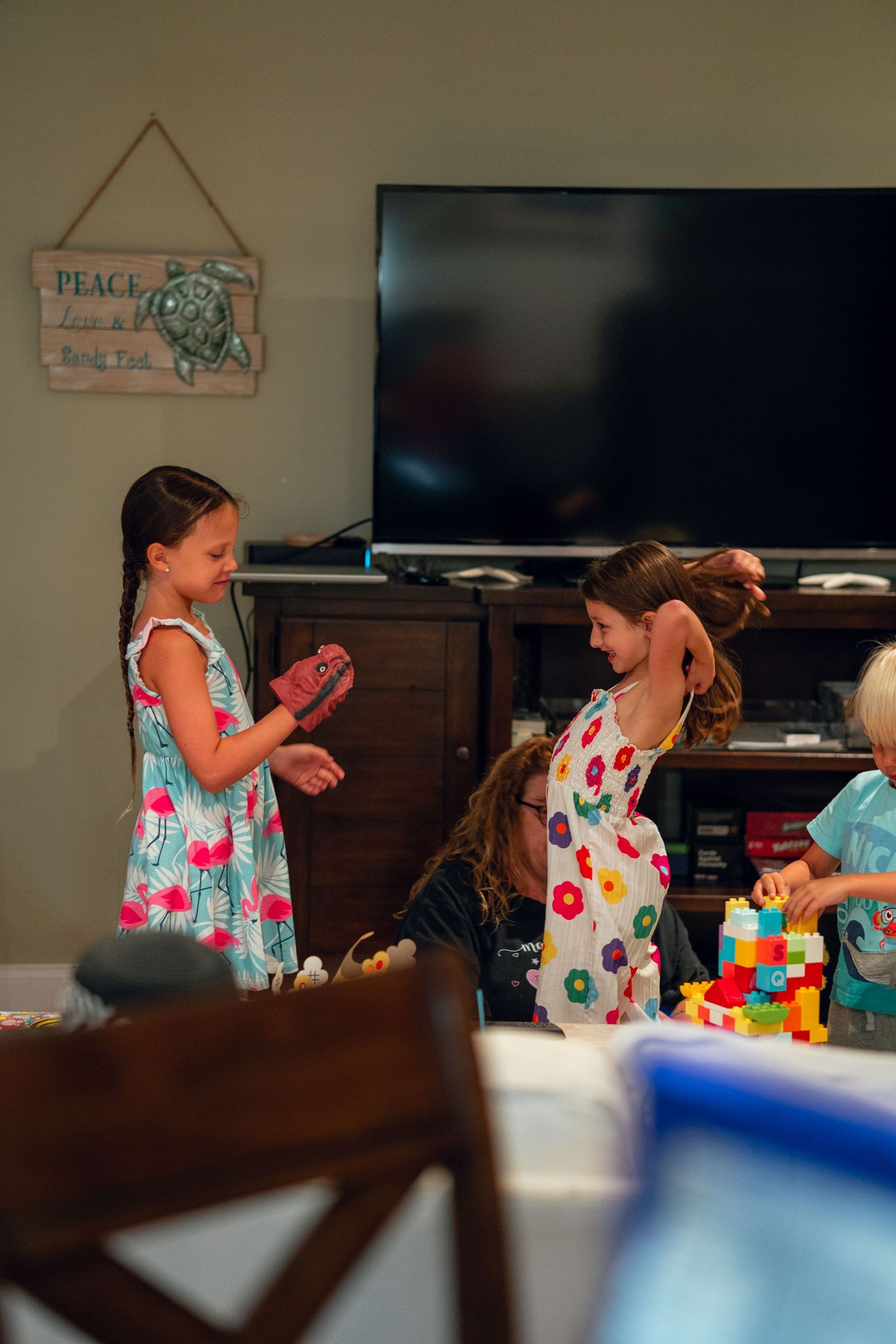 Two young girls in floral dresses playing and dancing in a living room with a TV cabinet.