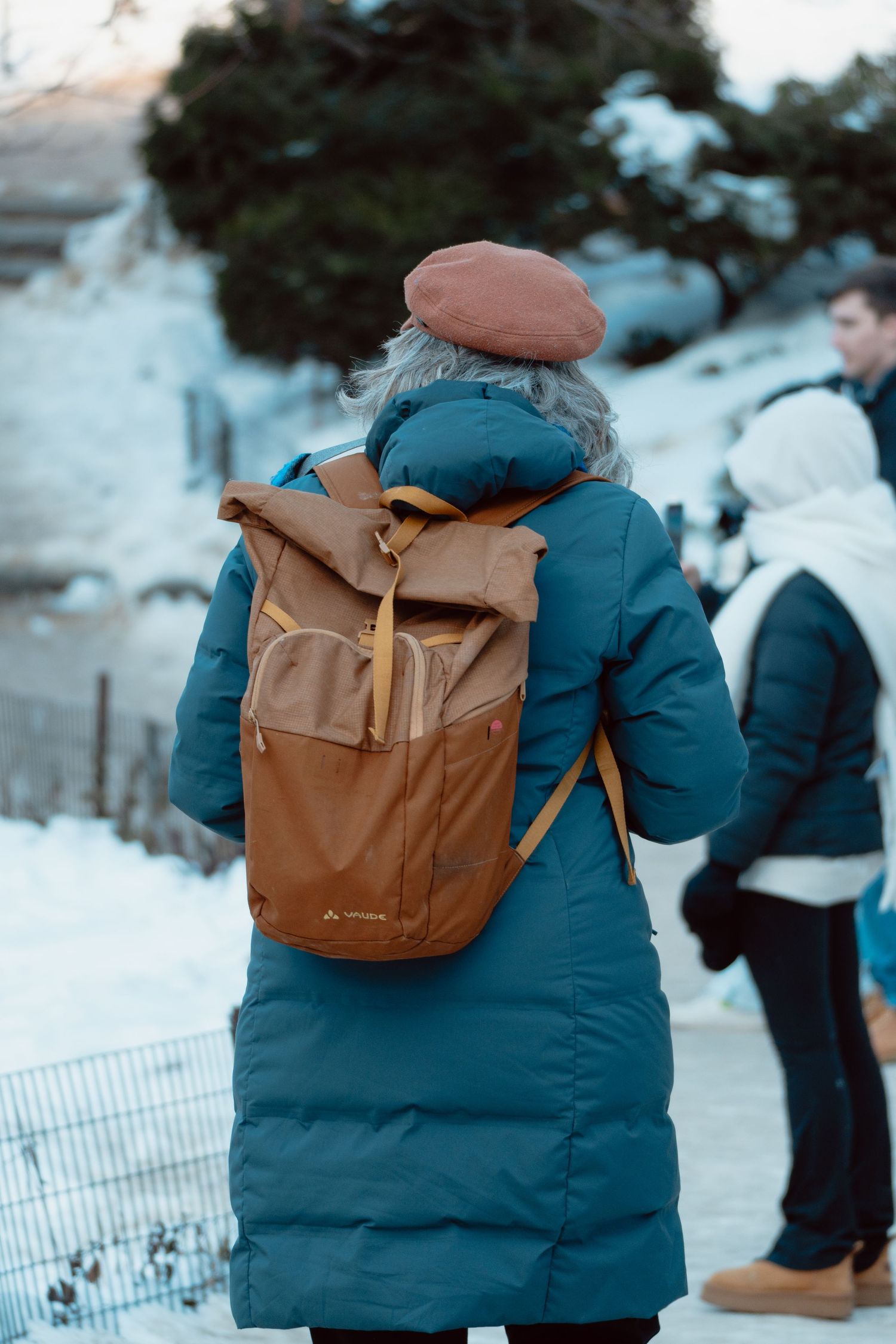 Woman in teal puffer coat and brown beret with backpack standing in snowy landscape.