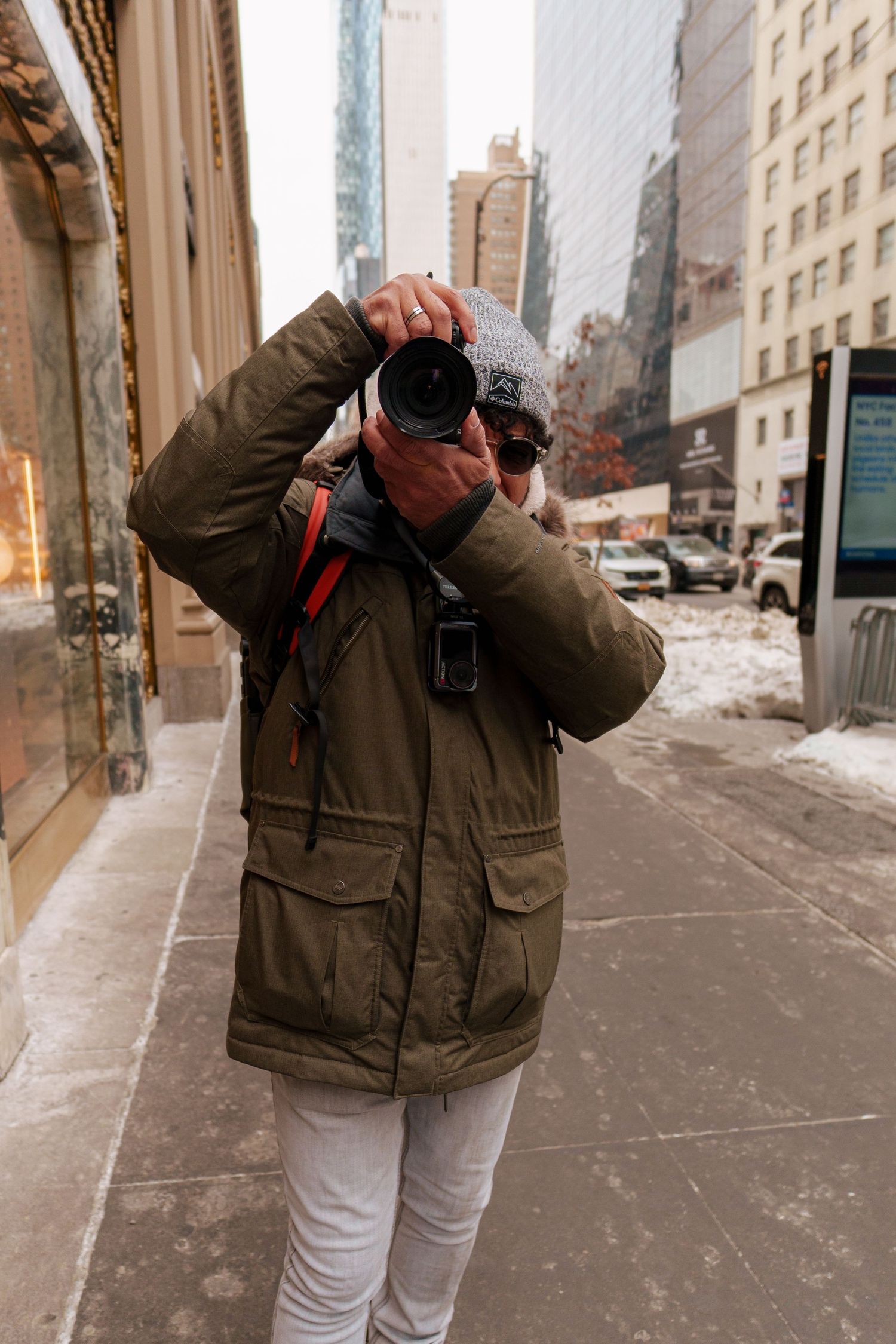 A photographer in a green winter jacket and gray beanie photographs on a snowy New York City street.
