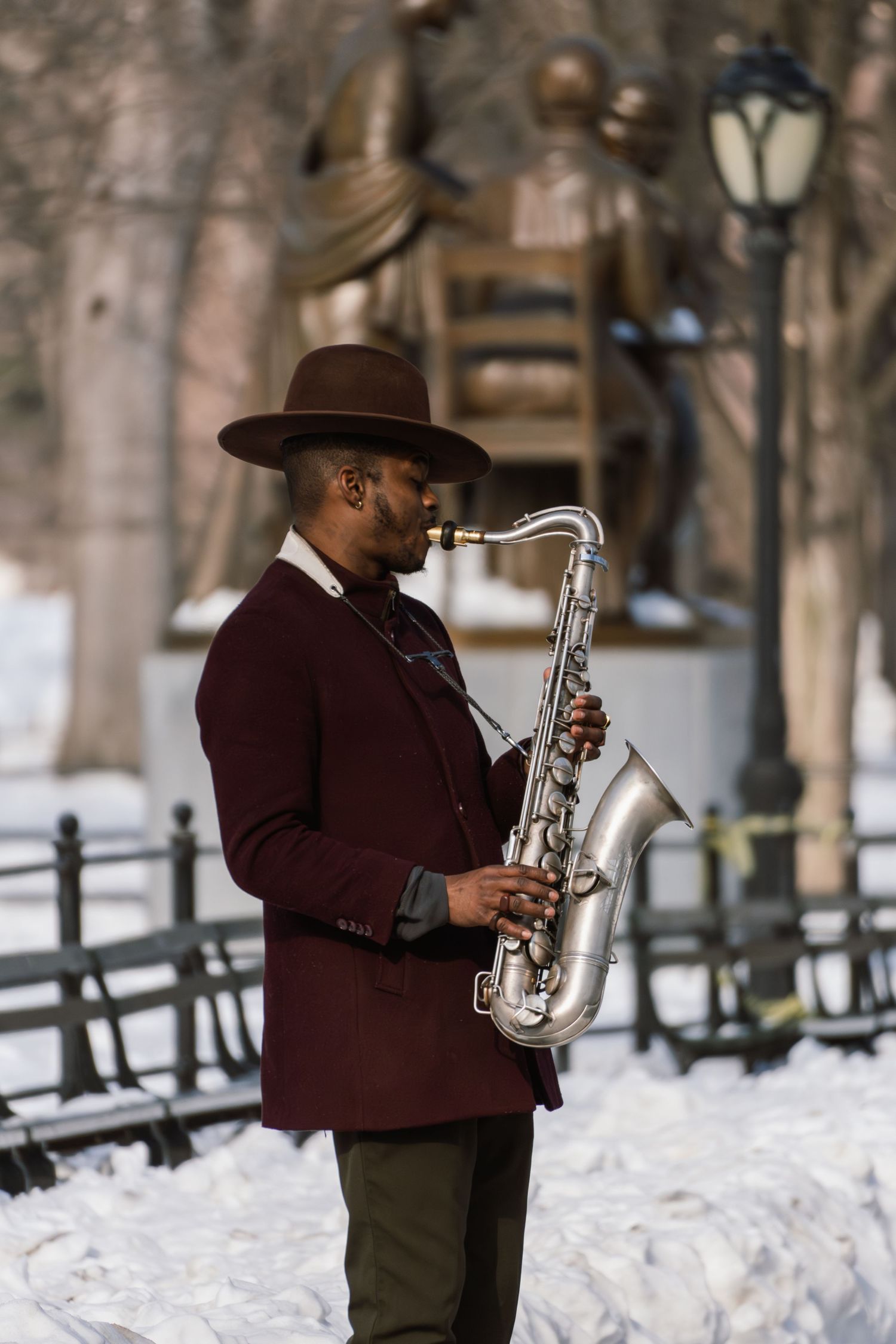 A stylish jazz musician in a burgundy jacket and wide-brim hat plays saxophone in a snow-covered park.