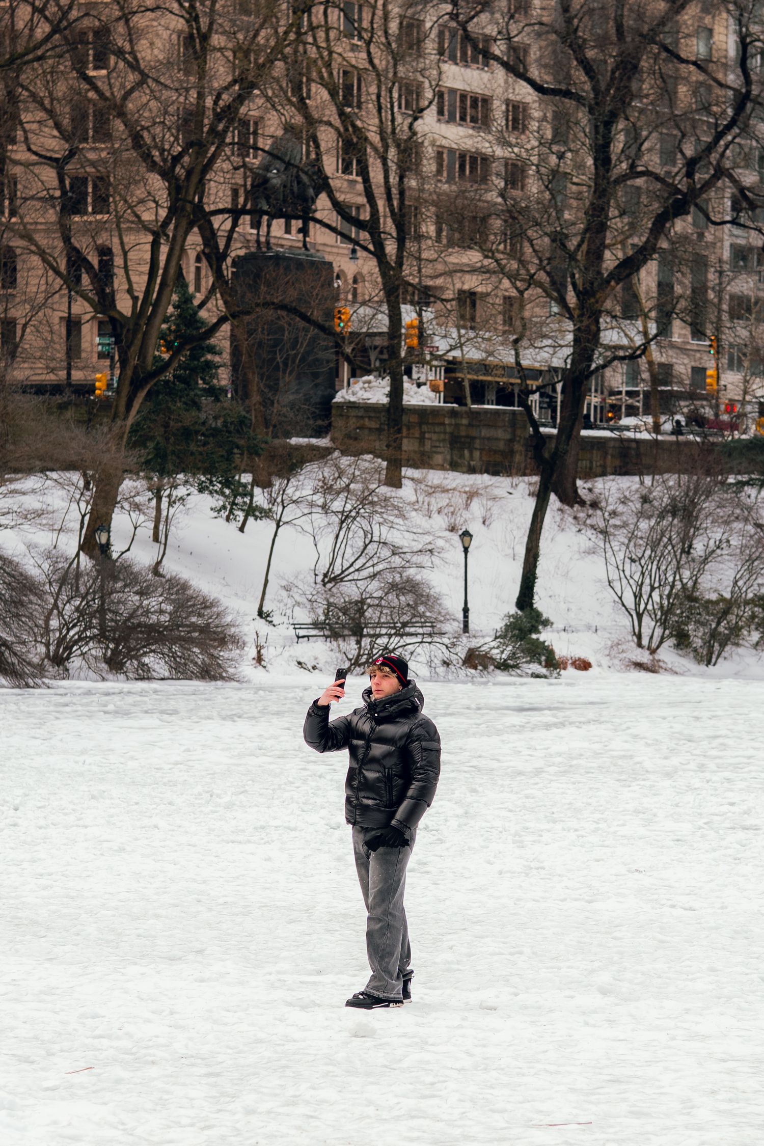 Person in black puffer jacket taking smartphone photos on a frozen snowy Central Park lake.