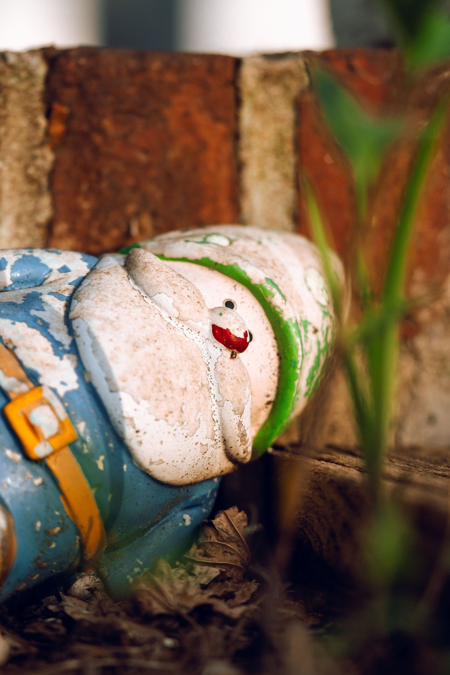 Close-up of a faded ceramic garden gnome lying on its back beside a brick wall and green plants.