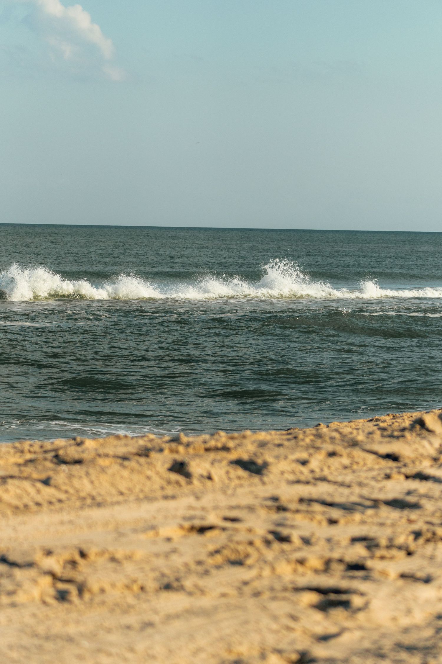 Multiple white-capped ocean waves crash along a sandy beach shoreline under a pale blue sky.