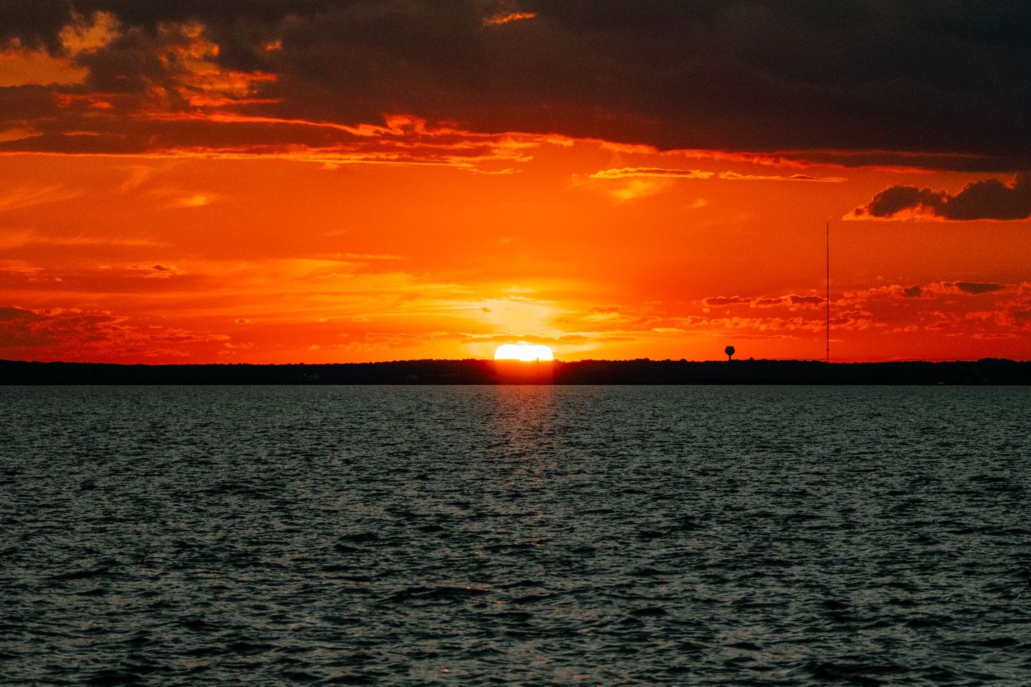 Dramatic fiery red and orange sunset over calm water with silhouetted distant treeline.