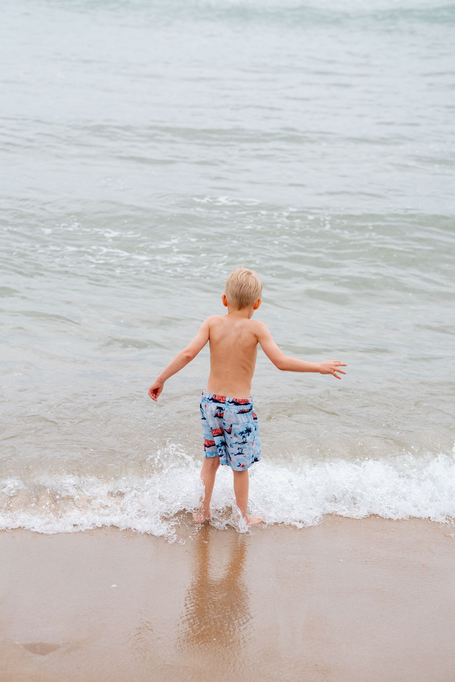 Young blond boy in swim trunks playing at ocean shoreline, splashing in gentle waves on sandy beach.