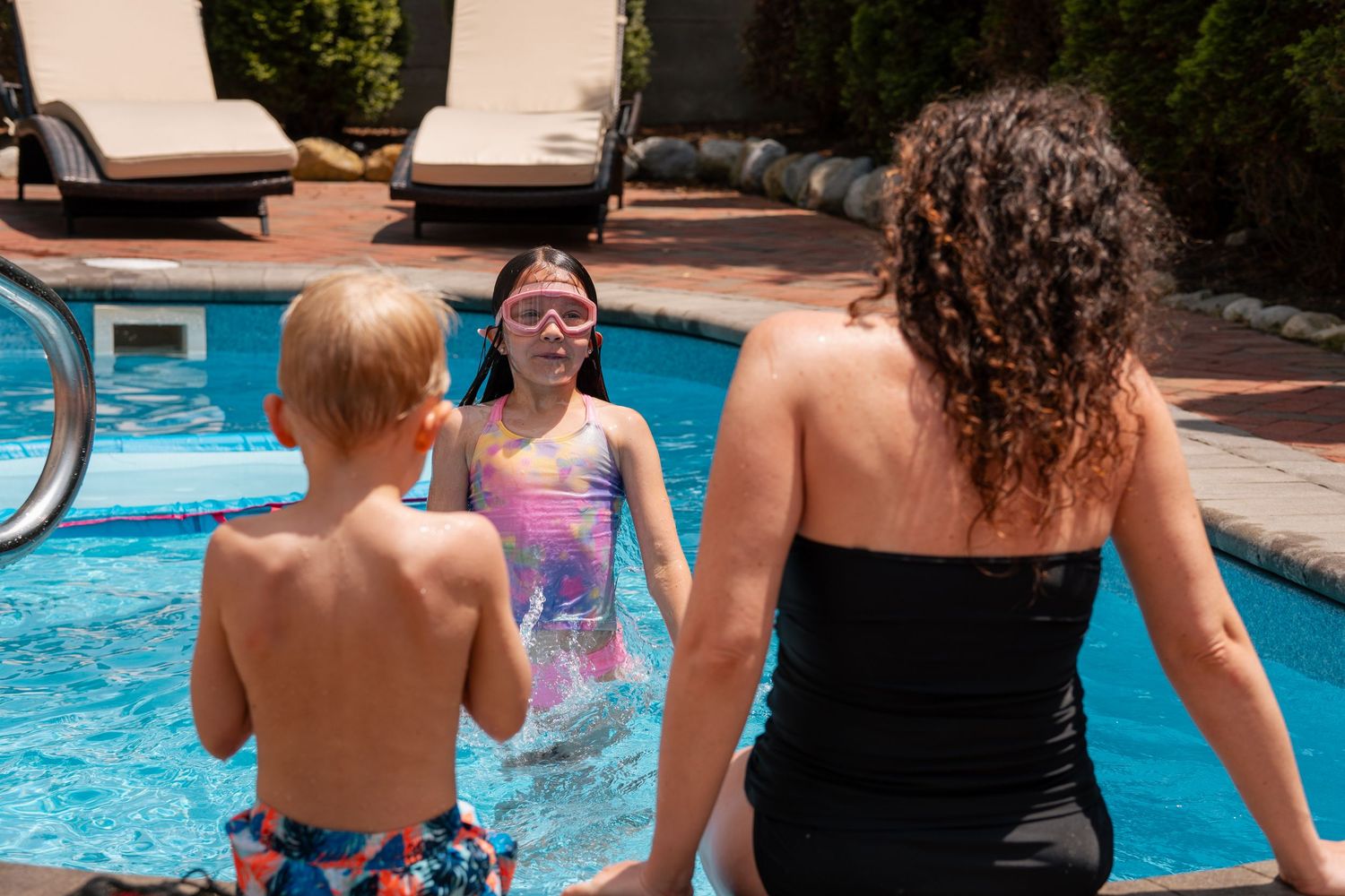 Woman and two children enjoying a sunny outdoor swimming pool, girl wearing pink goggles in the water.