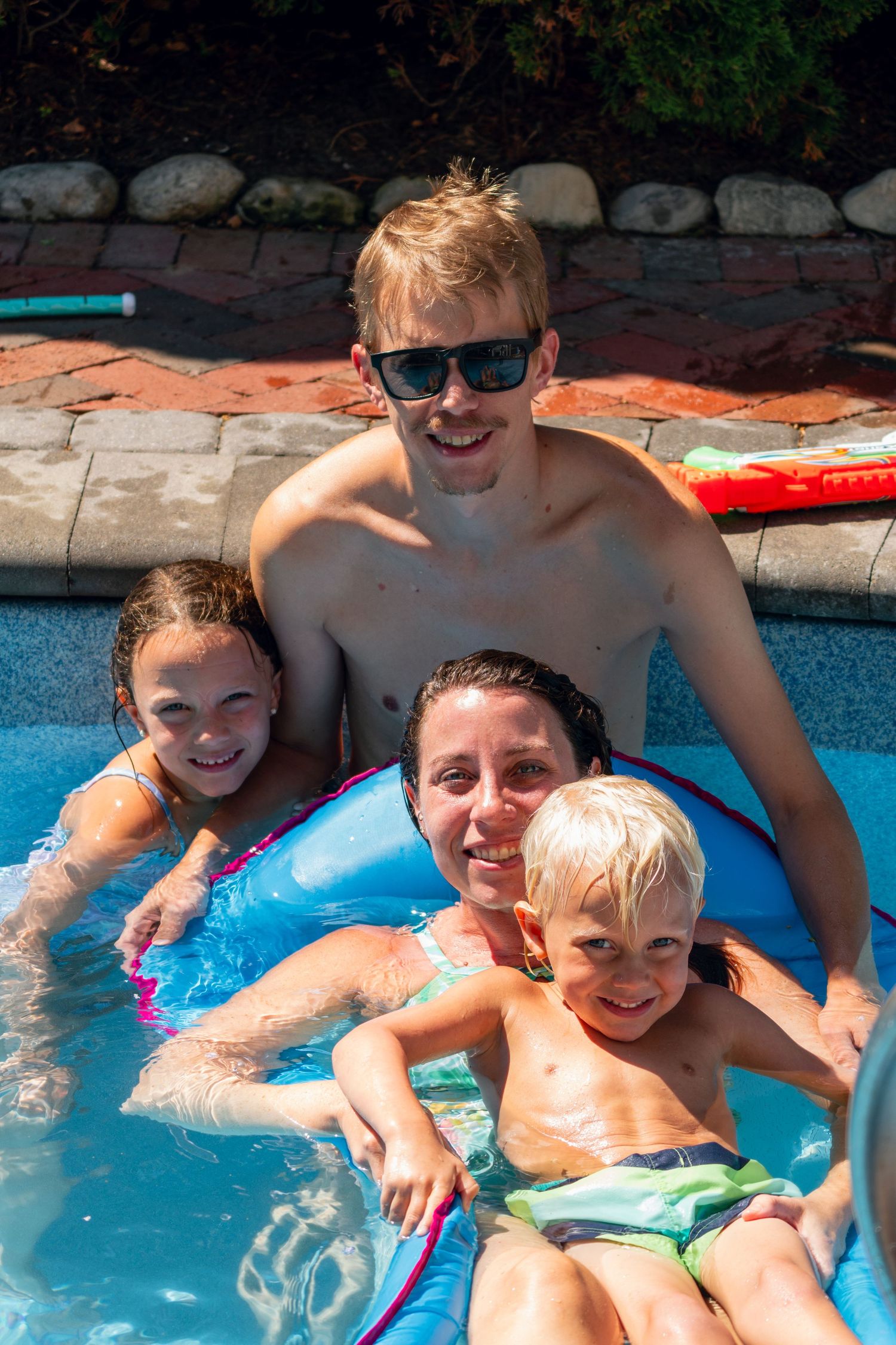 Smiling family of four posing together in a sunny backyard swimming pool with a float.