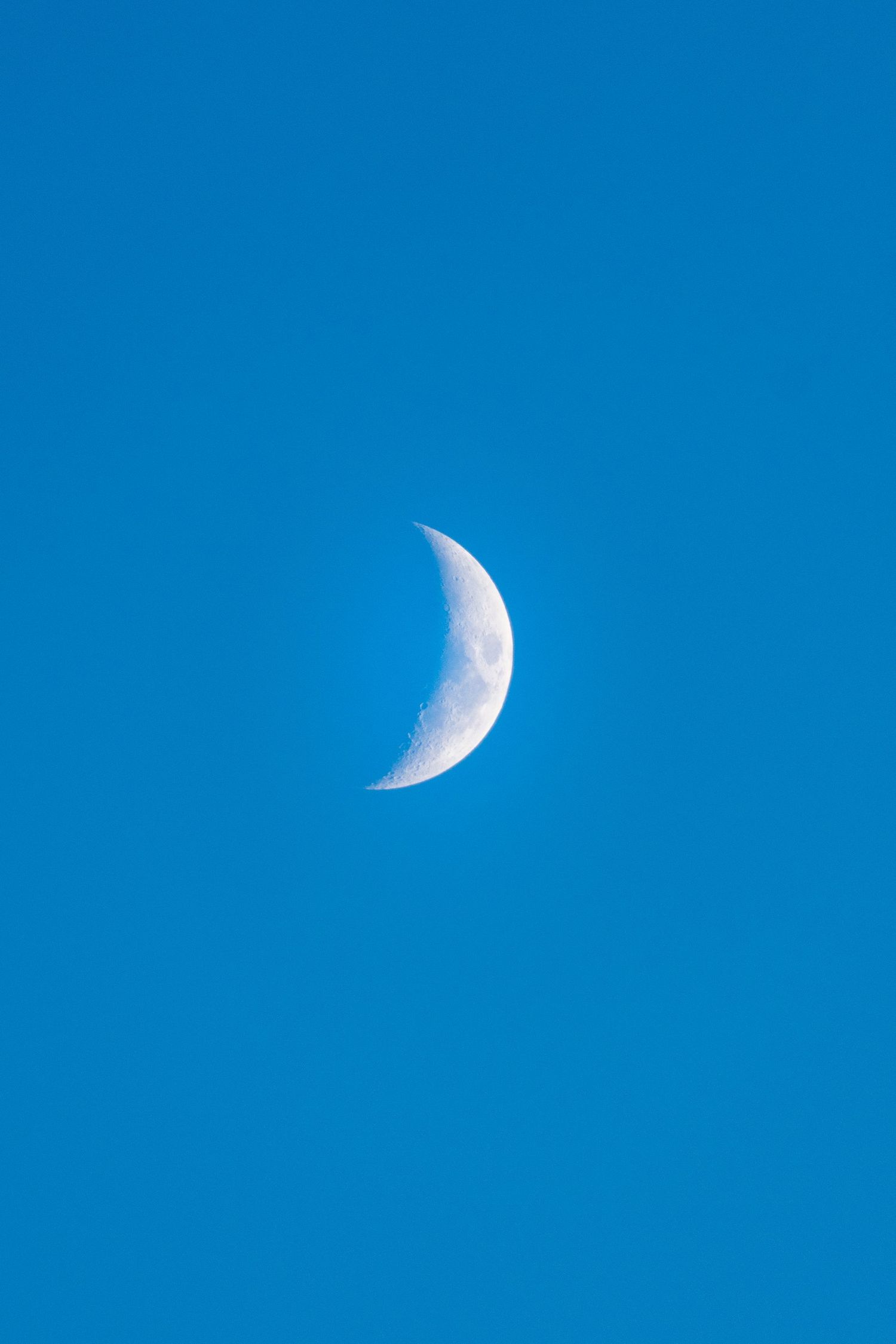 A crescent moon glowing white against a clear deep blue daytime sky.
