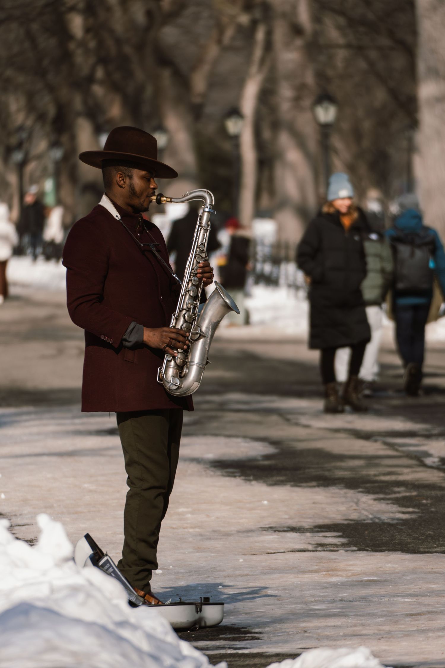 A street musician in a brown hat plays saxophone on a snowy Central Park path in winter.
