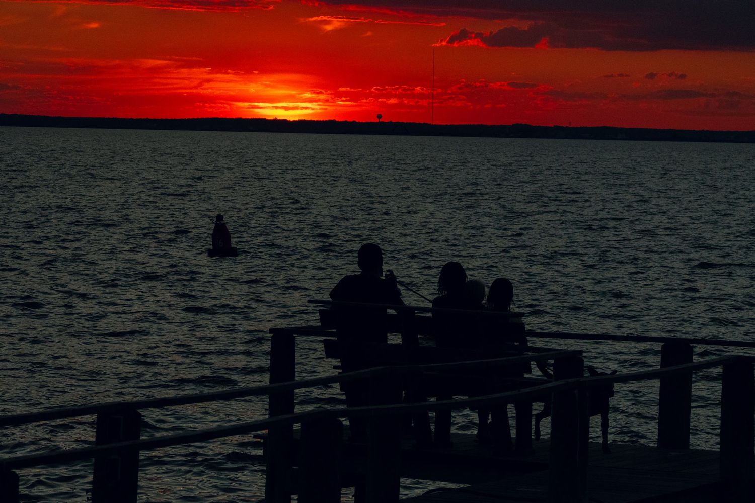 Three silhouetted figures on a waterfront dock beneath a deep red dramatic sunset sky.