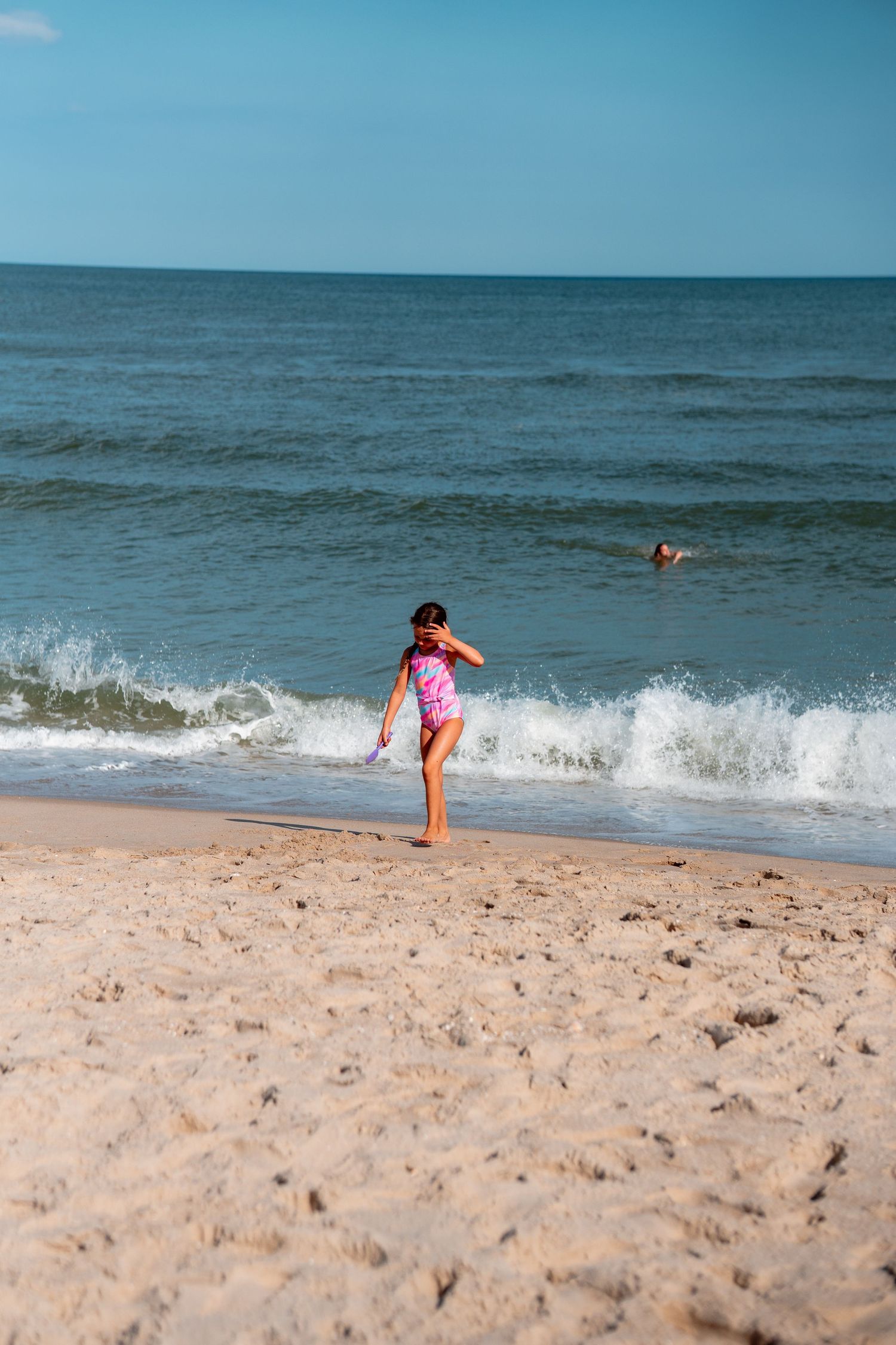 Young girl in pink swimsuit playing at the ocean shoreline with waves on a sunny day.