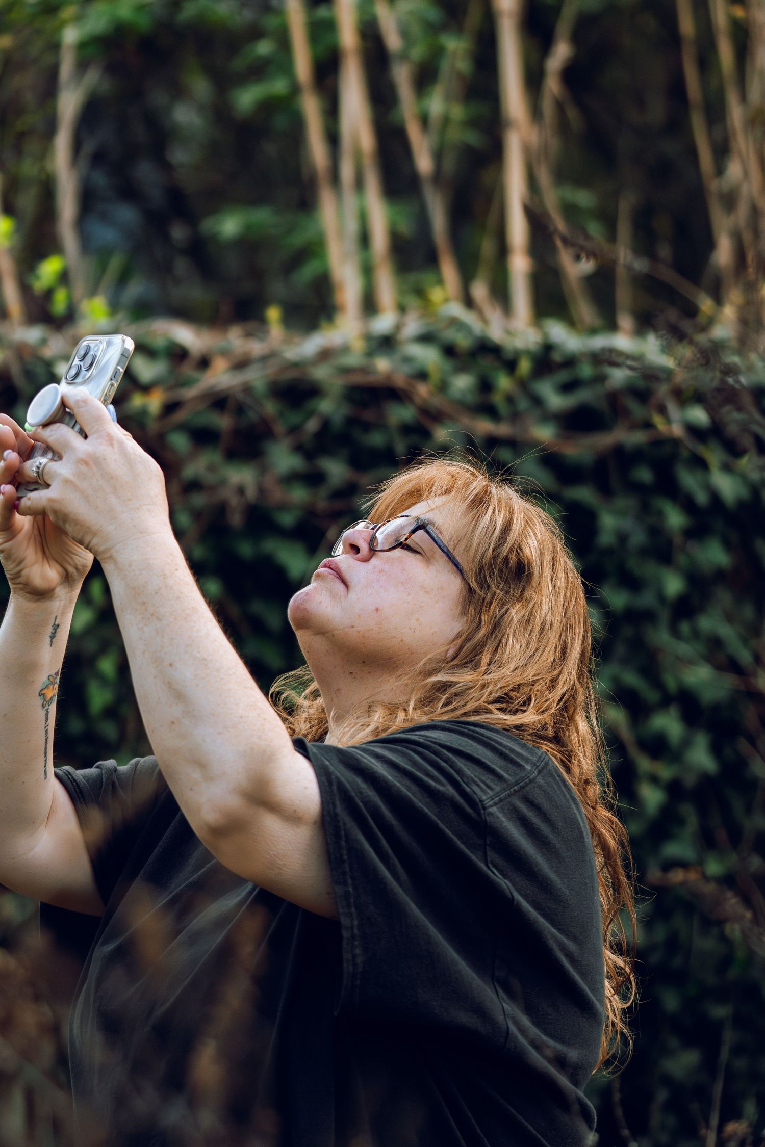 A woman with red hair and glasses holds a camera up to photograph something above her in a lush green garden.