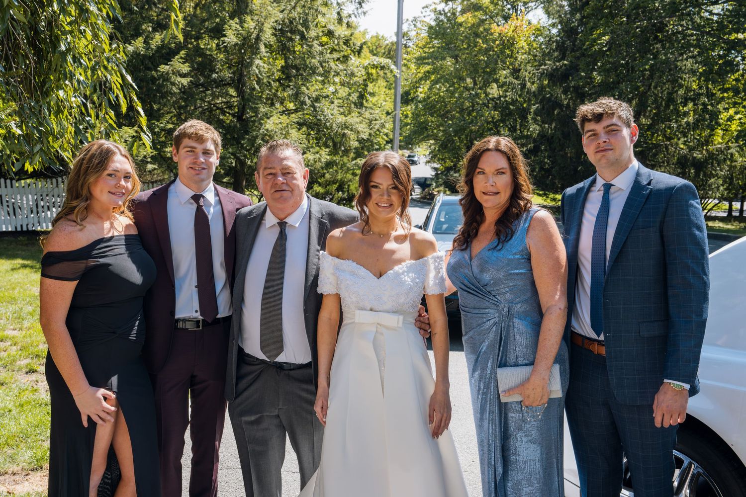A bride in white dress poses with five formally dressed guests outdoors at a sunny wedding celebration.