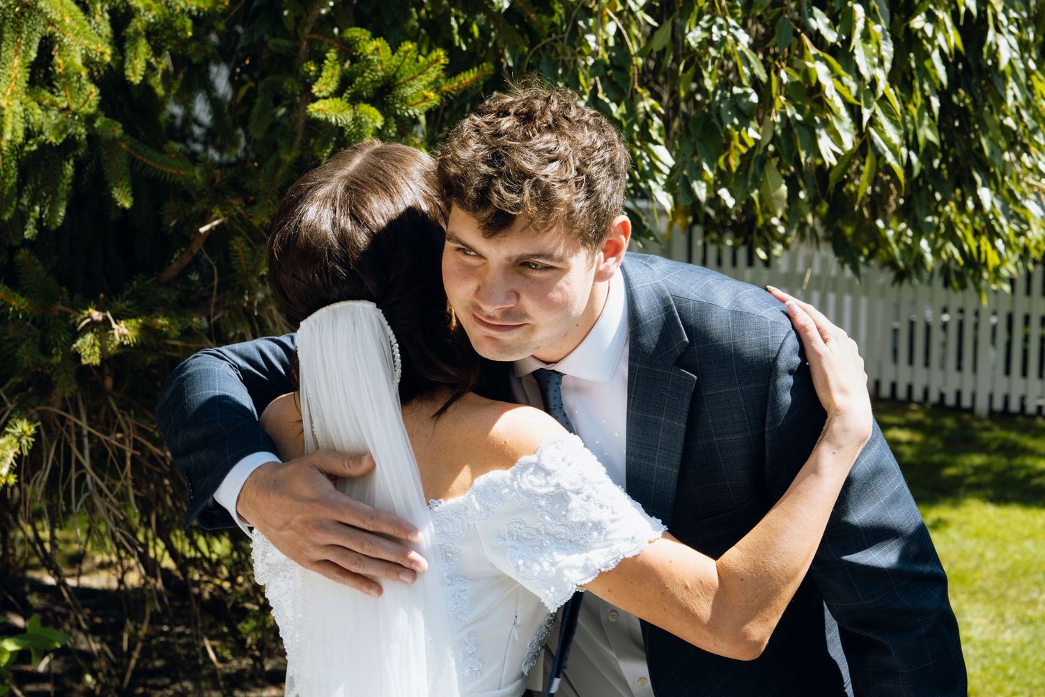 A groom in a navy suit embraces a bride in a white lace dress outdoors near green trees and a white fence.