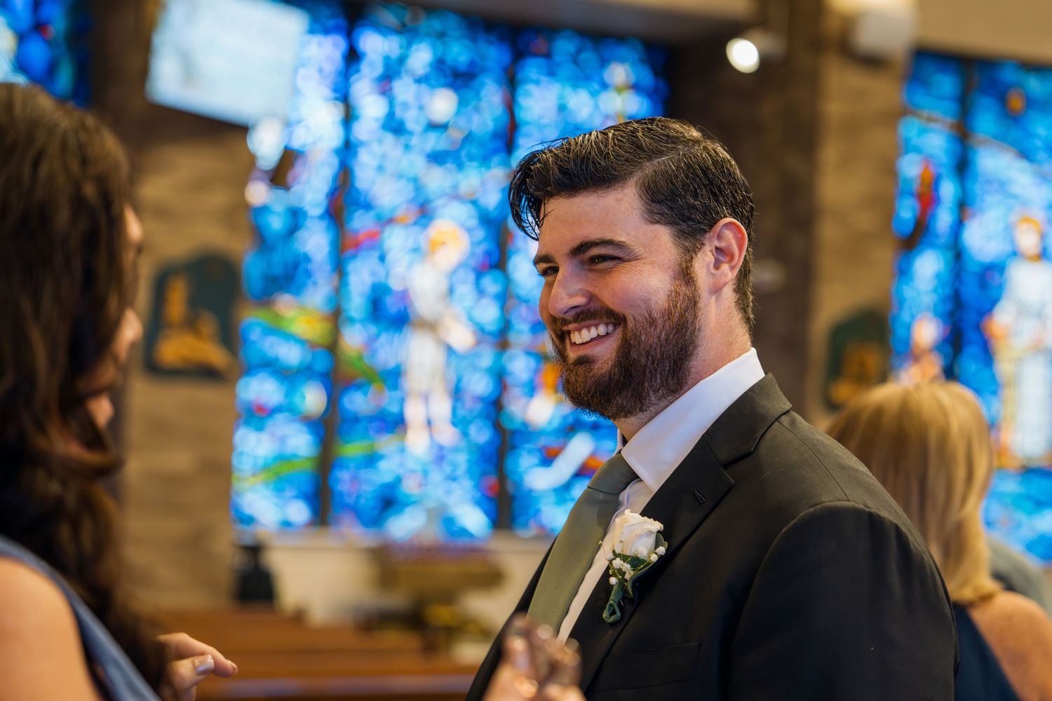 Smiling groom with beard in suit at church wedding ceremony with colorful stained glass windows in background.