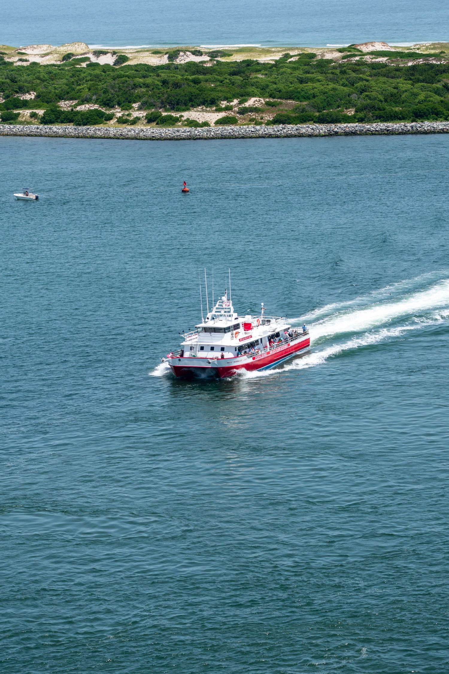 Red and white passenger ferry boat cruising through blue ocean waters near a sandy island coastline.