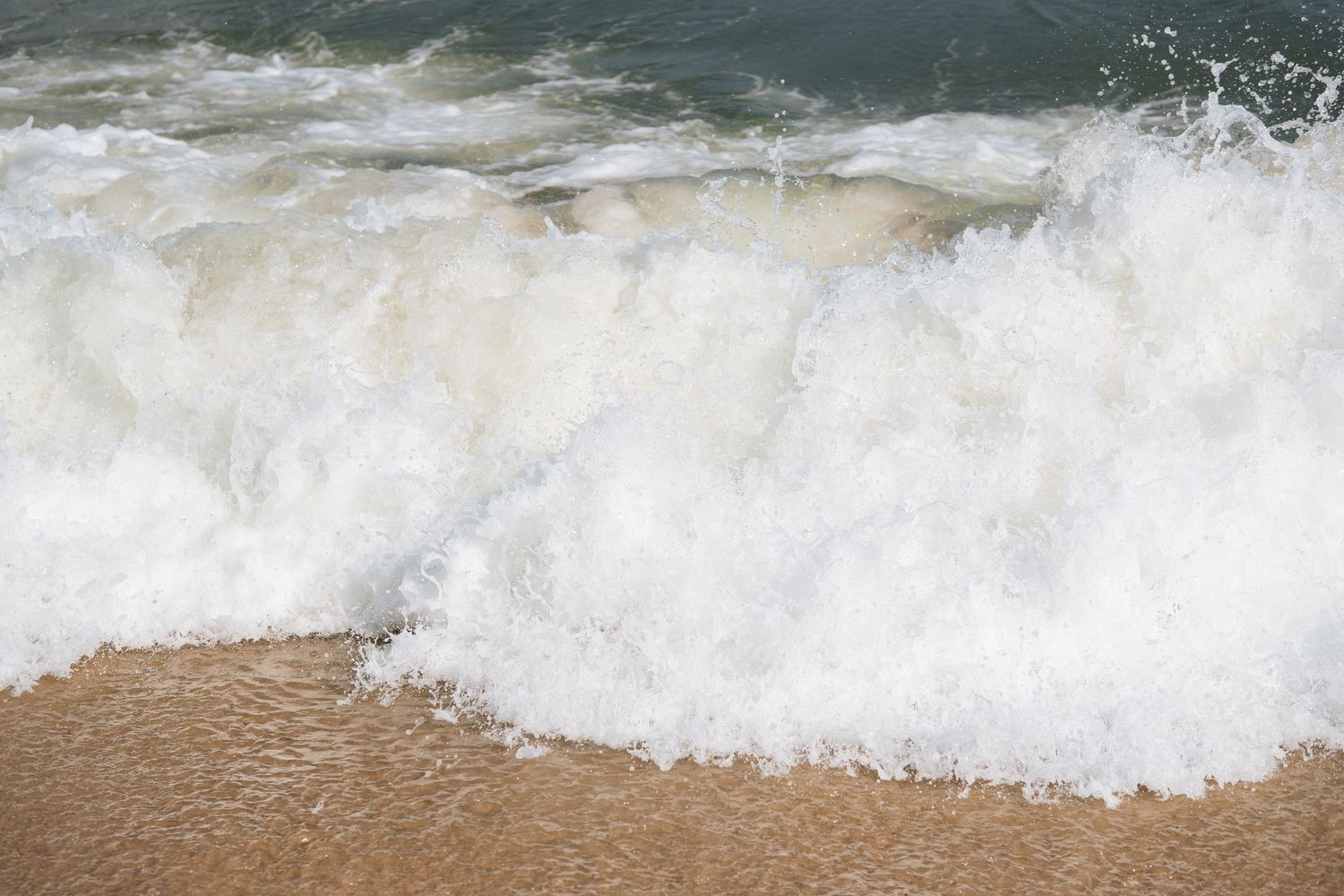 Powerful white foamy ocean wave crashing onto a sandy beach shore close-up view.