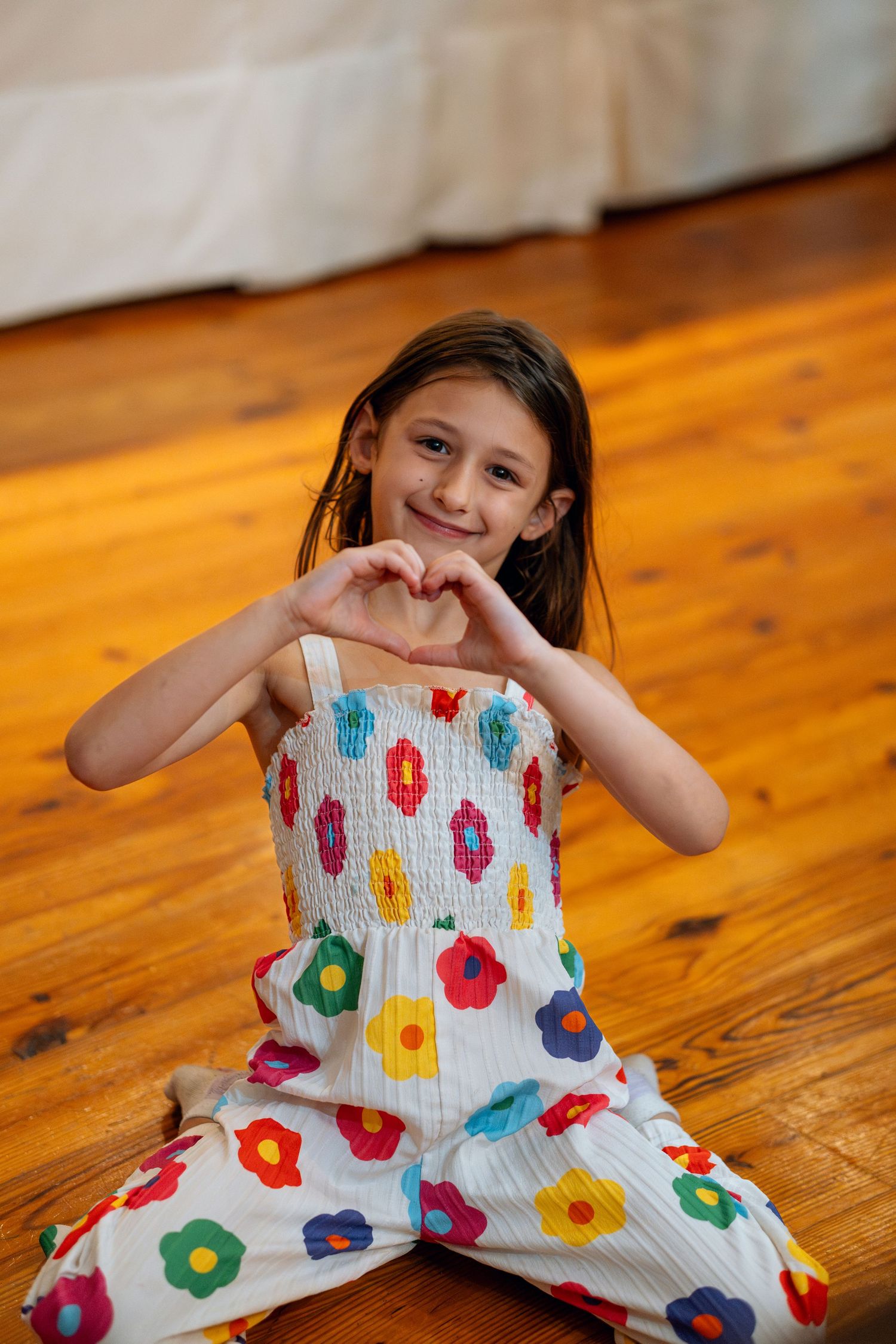 Young girl in a colorful floral jumpsuit making a heart shape with her hands on a hardwood floor.