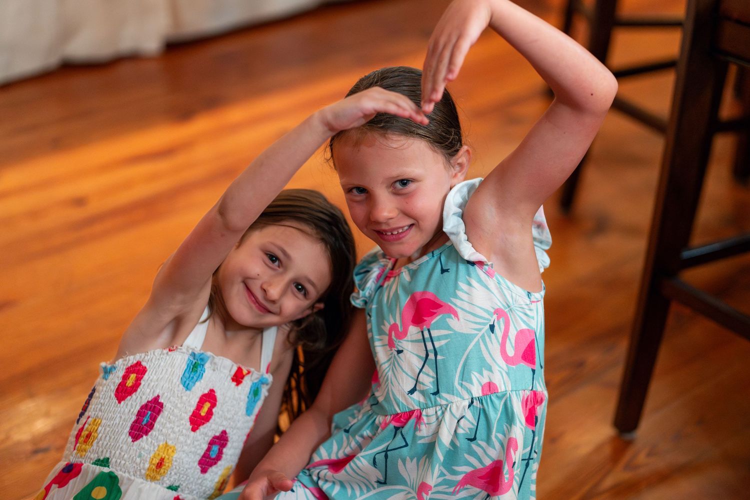 Two cheerful young girls posing together on a hardwood floor, raising their arms playfully.