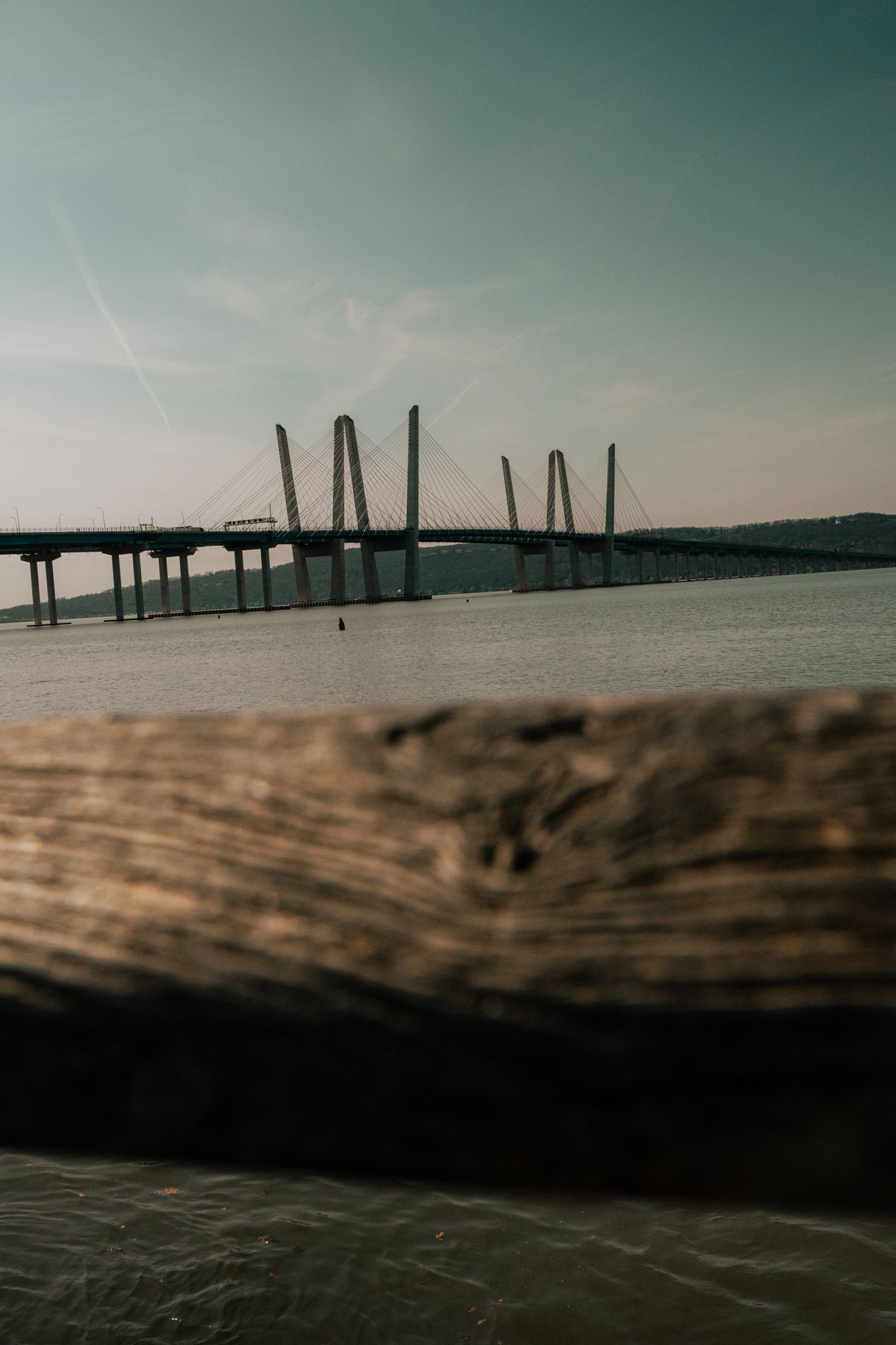 A cable-stayed bridge spans a wide river, viewed from behind a weathered wooden log in the foreground.