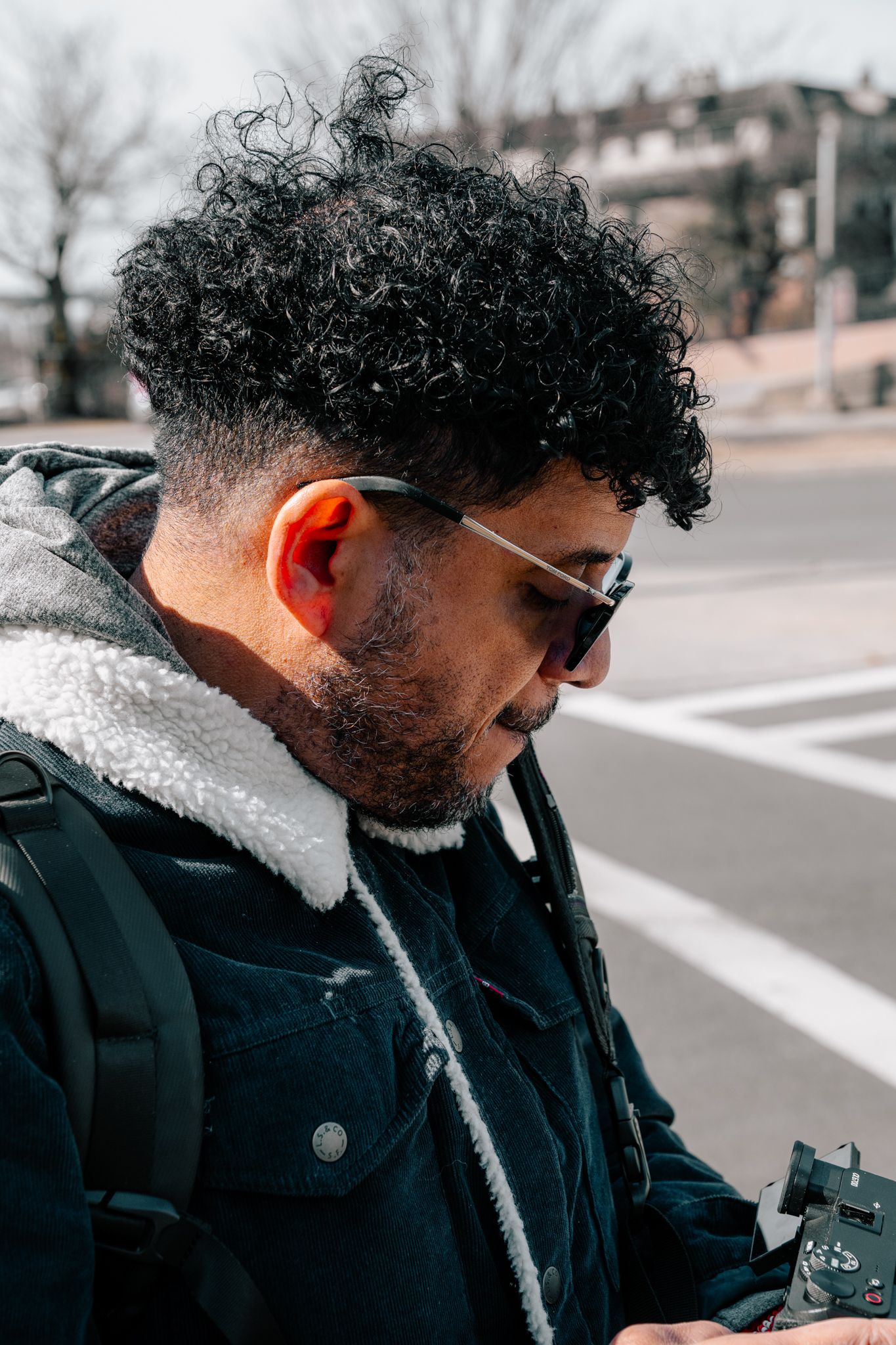 Close-up portrait of a curly-haired bearded man with sunglasses reviewing photos on a camera outdoors.