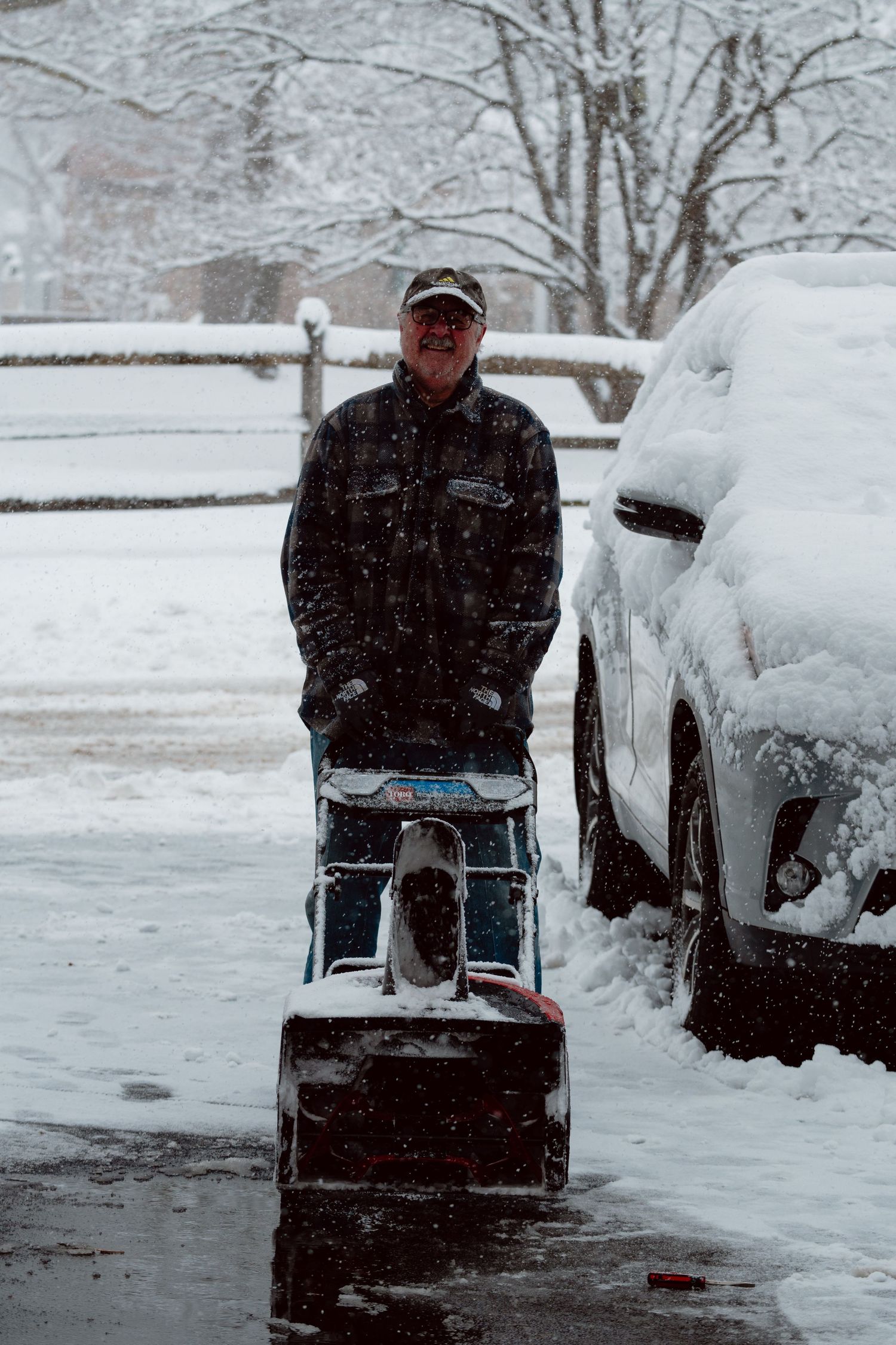 Smiling man operating a snowblower near a snow-covered car during a heavy winter snowfall.