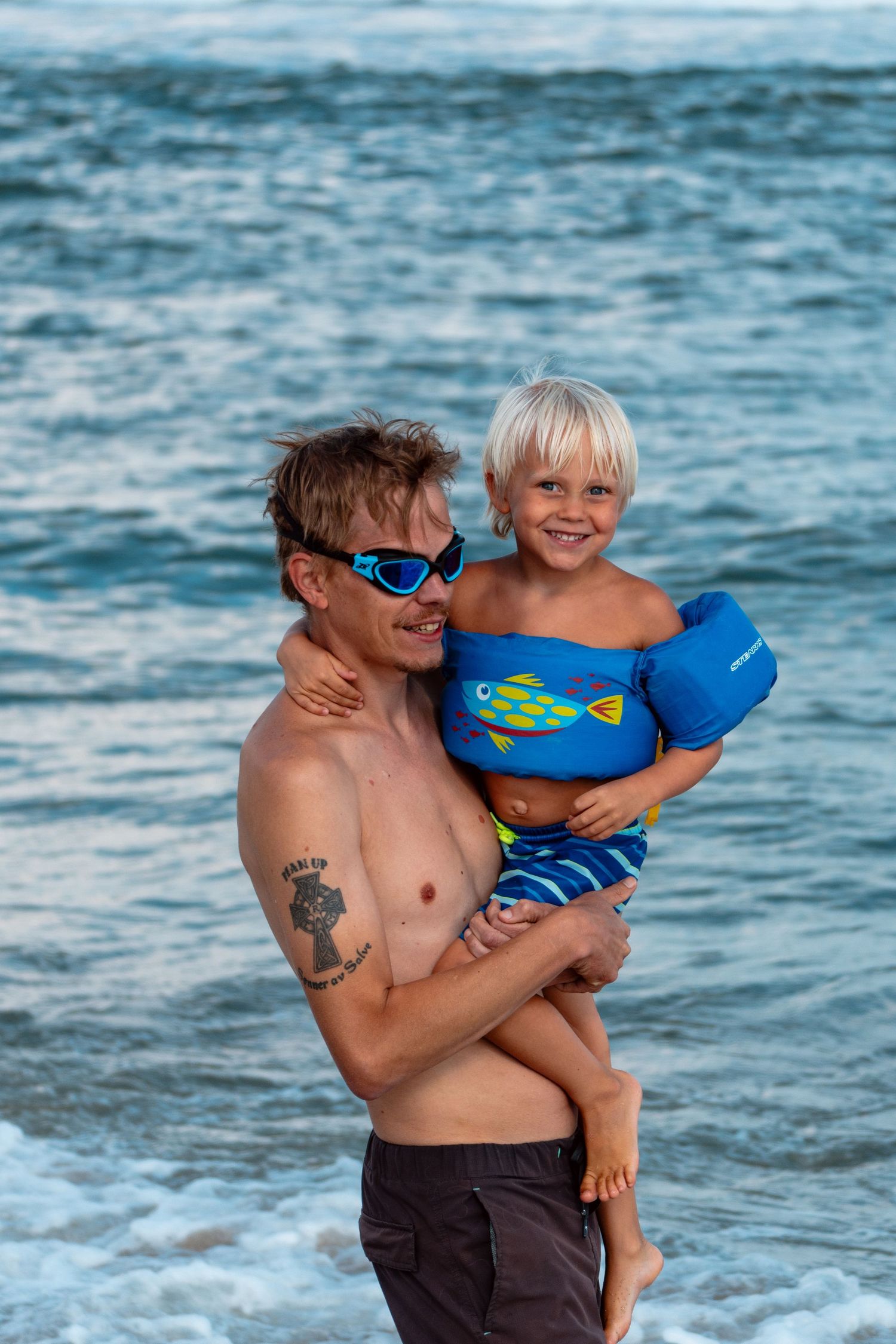Smiling father holding young blonde boy in blue floaties at the beach.