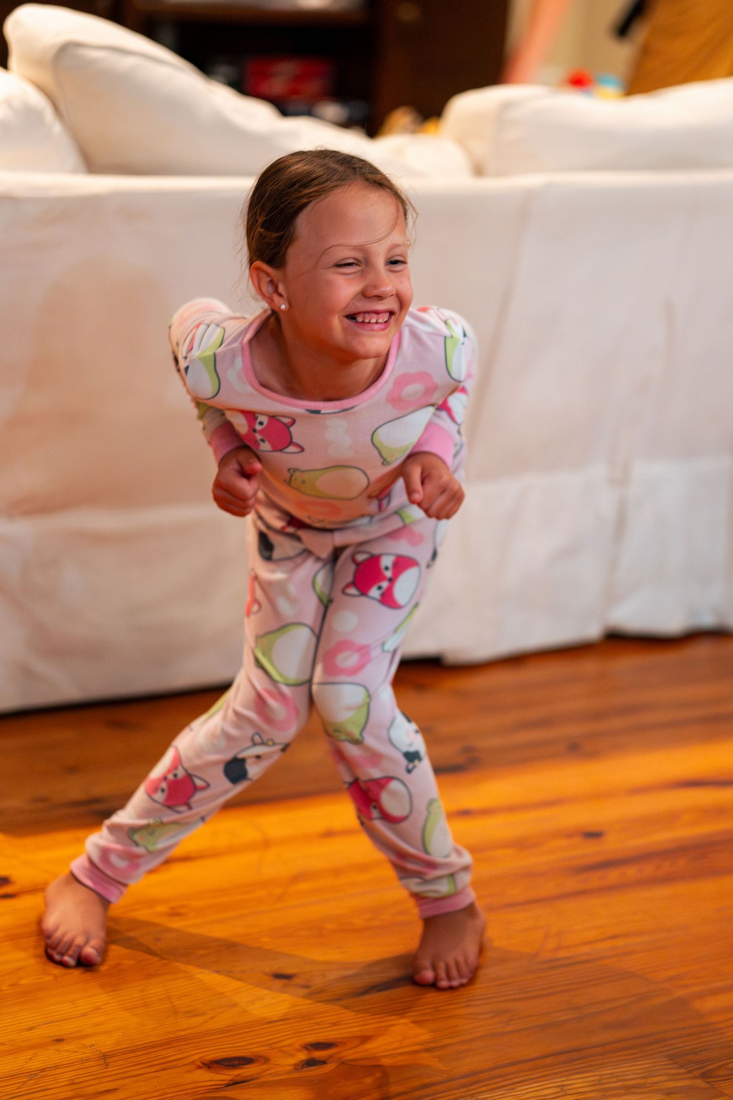 A laughing young girl in pink monster pajamas dances barefoot on a hardwood floor.