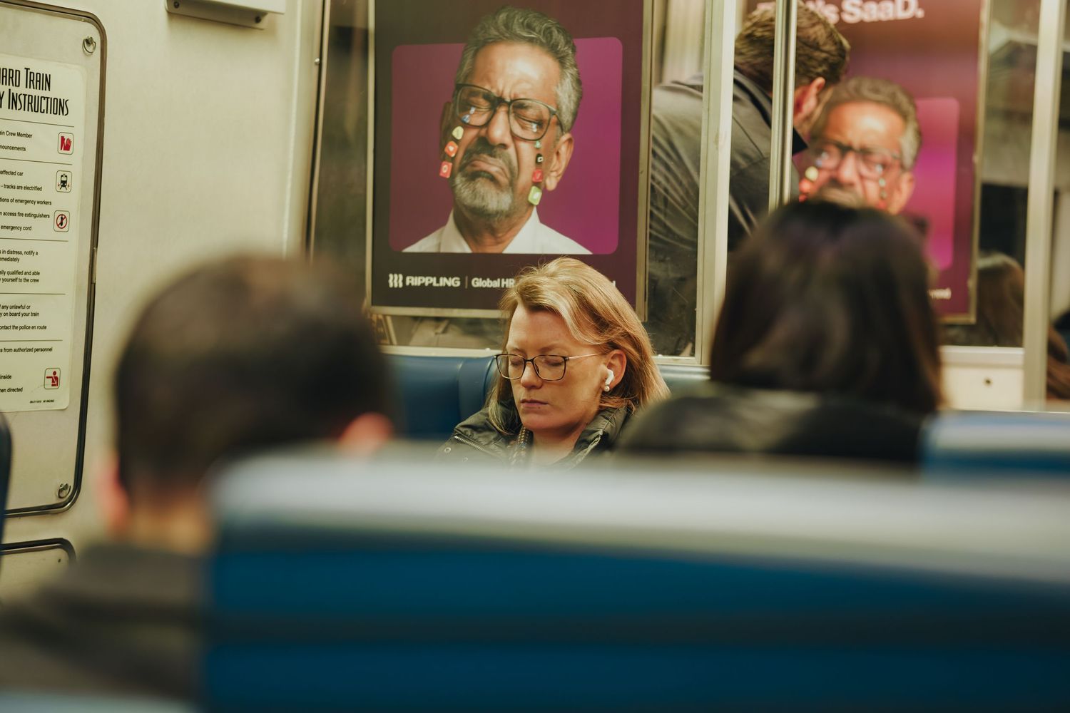 Woman with earbuds on subway train with advertisement poster behind her.