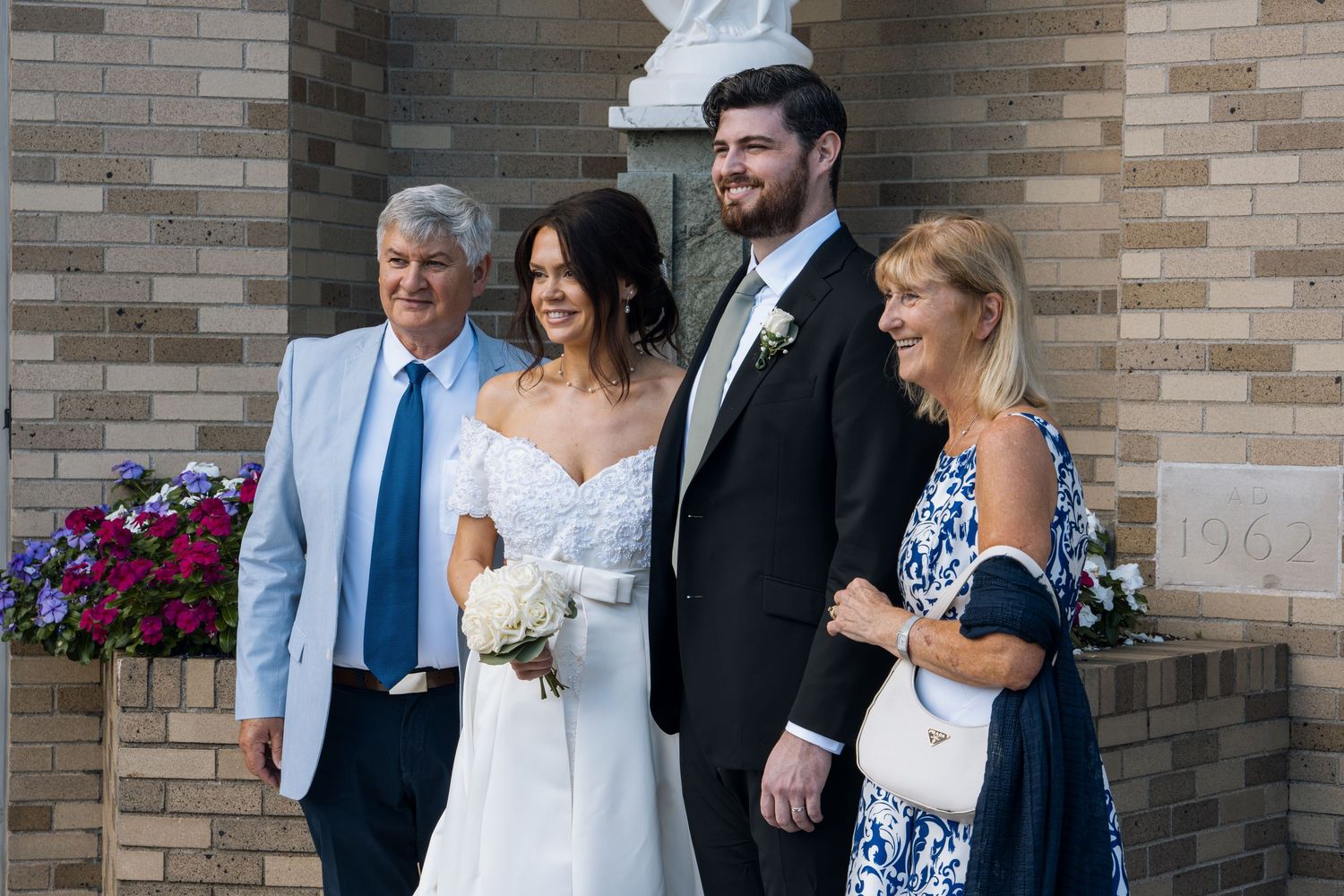 Wedding couple poses with family outside brick church, bride holding white bouquet in elegant off-shoulder gown.