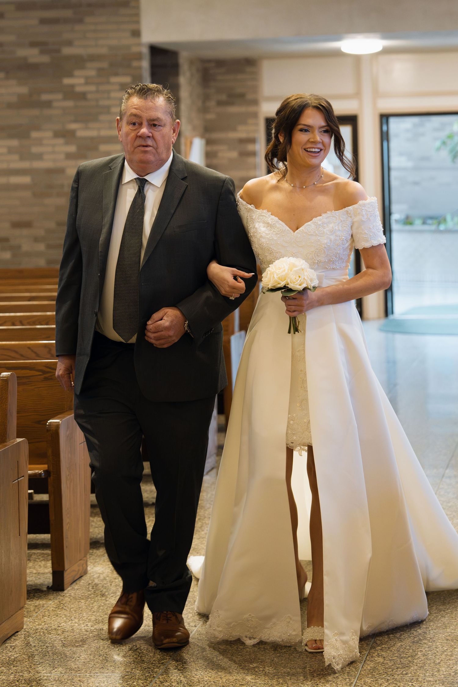 Bride in white off-shoulder gown walks down church aisle with older man in dark suit, carrying white bouquet.