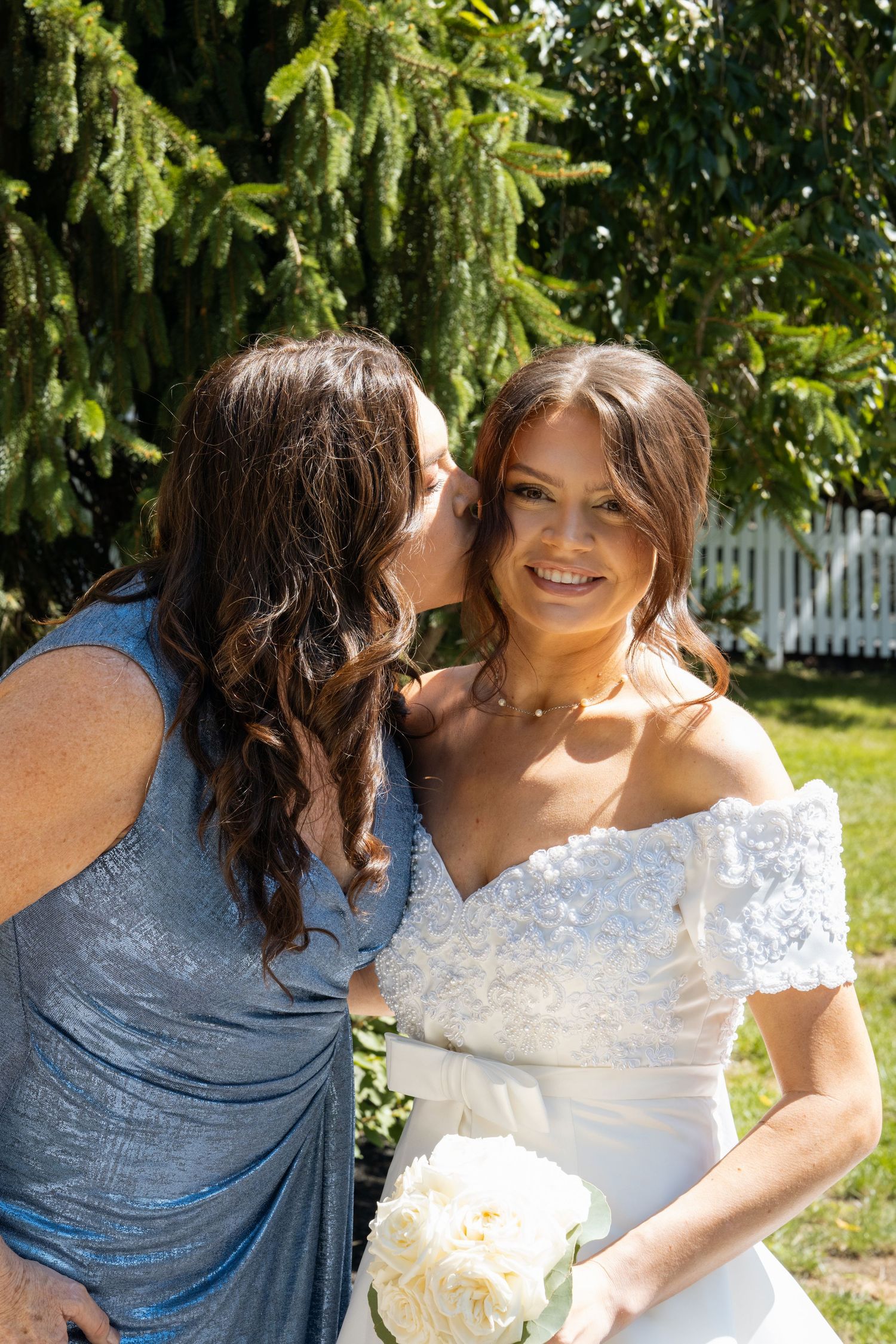 A bride in a white off-shoulder gown holds white peonies while receiving a kiss on the cheek from a woman in blue.