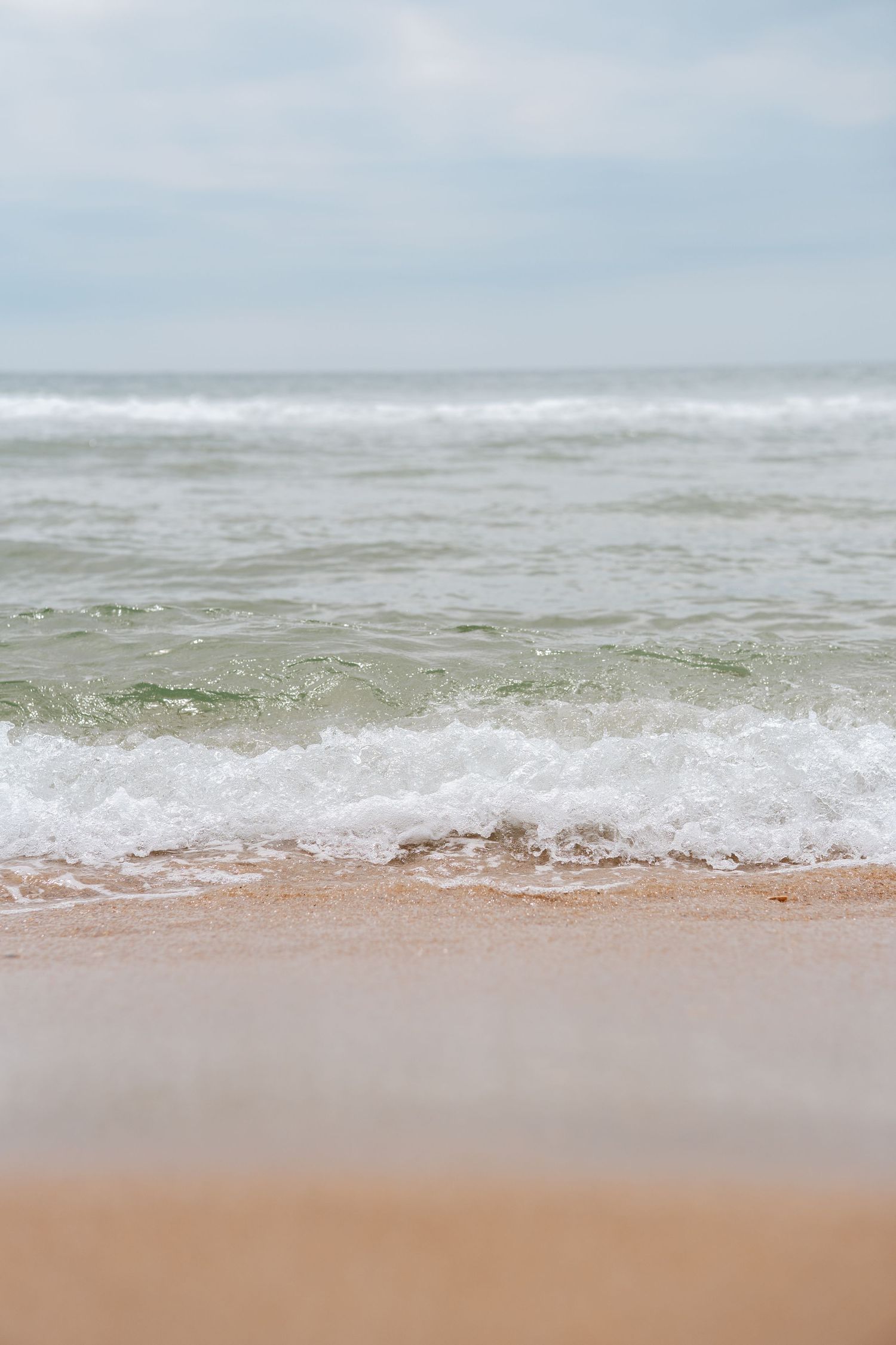 Gentle ocean waves washing onto a sandy beach shore under a cloudy sky, with turquoise water and white sea foam.